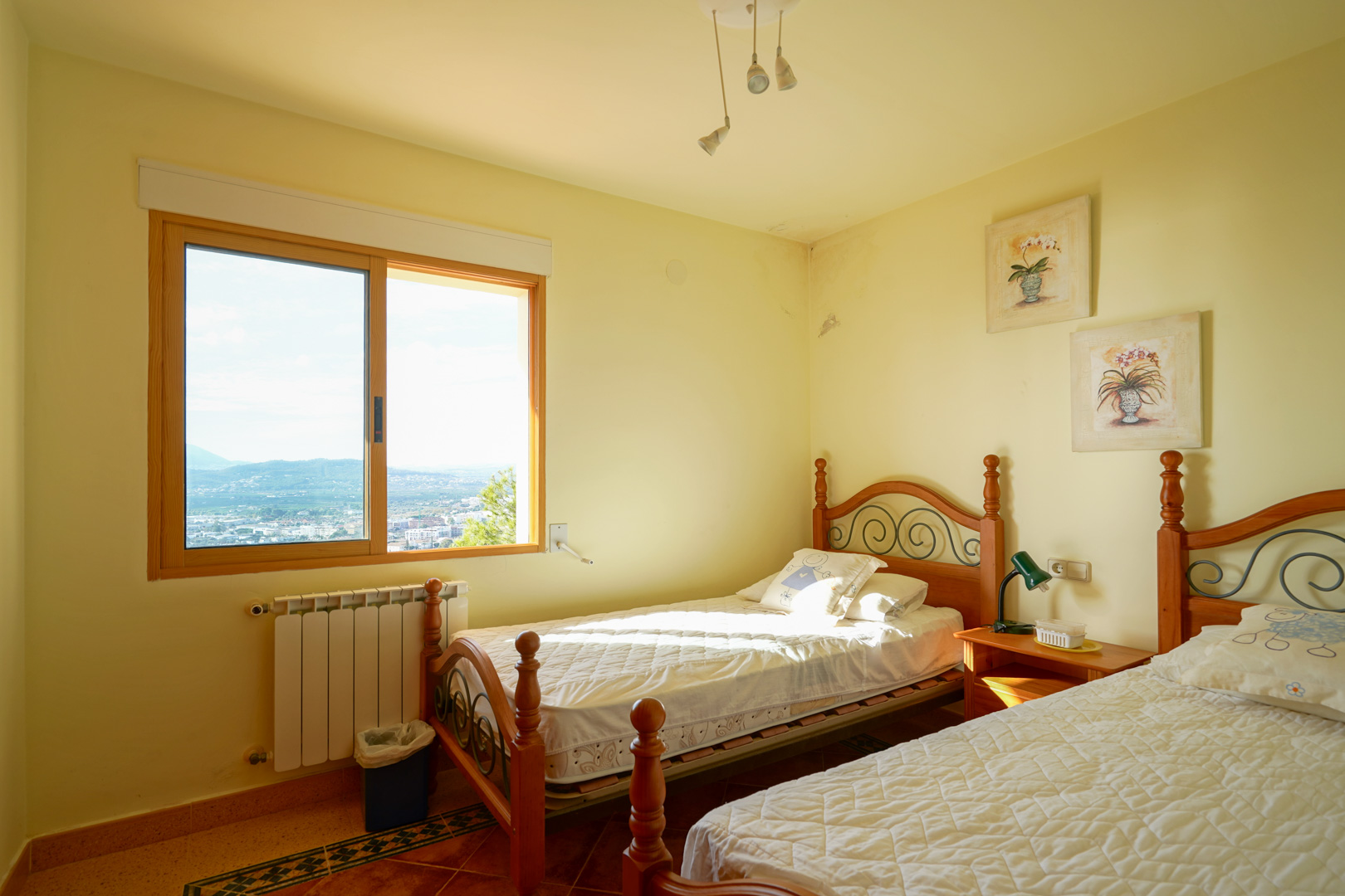 Zweibettzimmer mit Holz- und Schmiedeeisenbetten, Fliesenboden und großem Fenster mit weitem Blick über das Tal und die umliegende Berglandschaft.