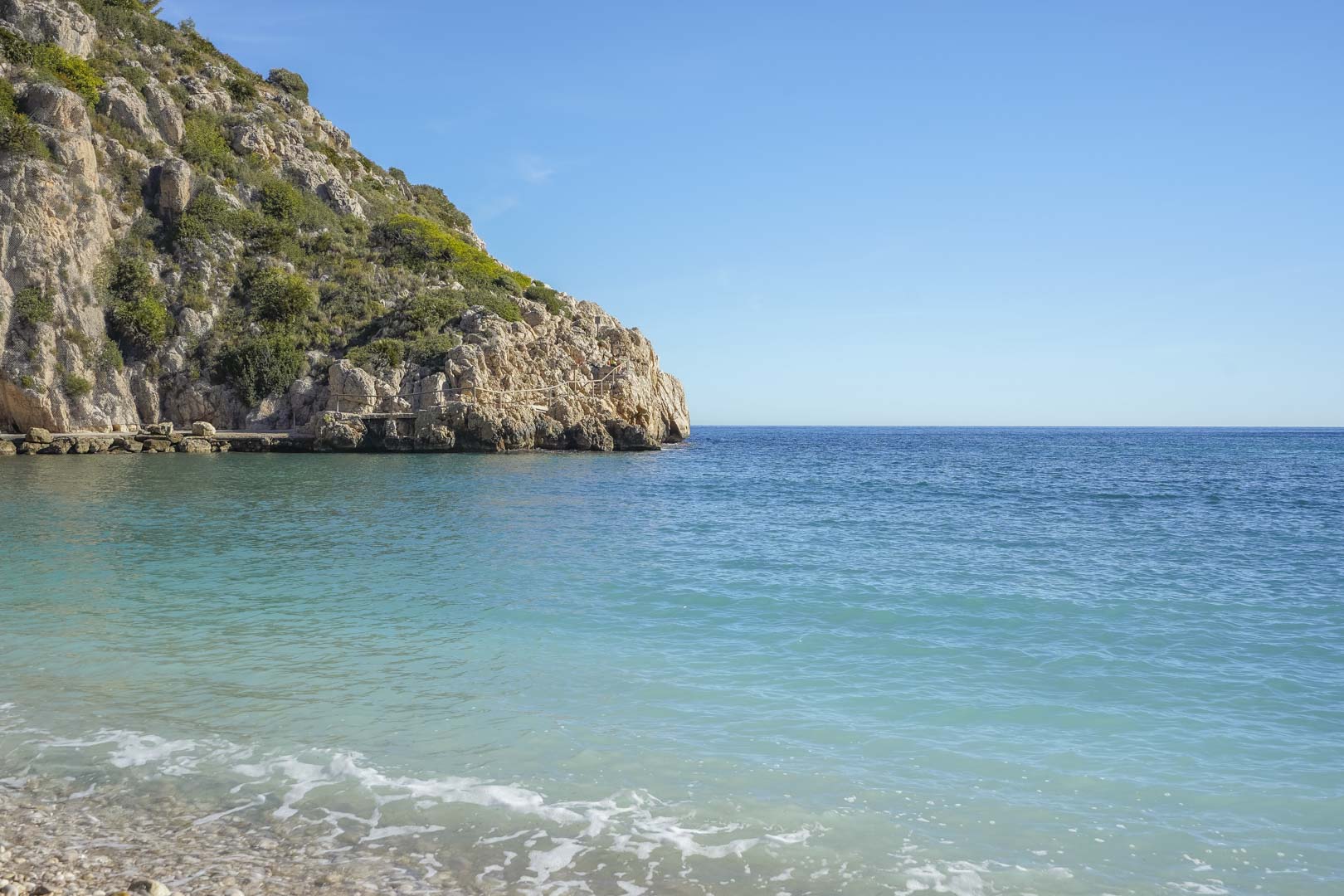 Blick auf das türkisfarbene Wasser der Cala Granadella mit Kieselstrand und markanten Kalksteinfelsen unter strahlend blauem Himmel.