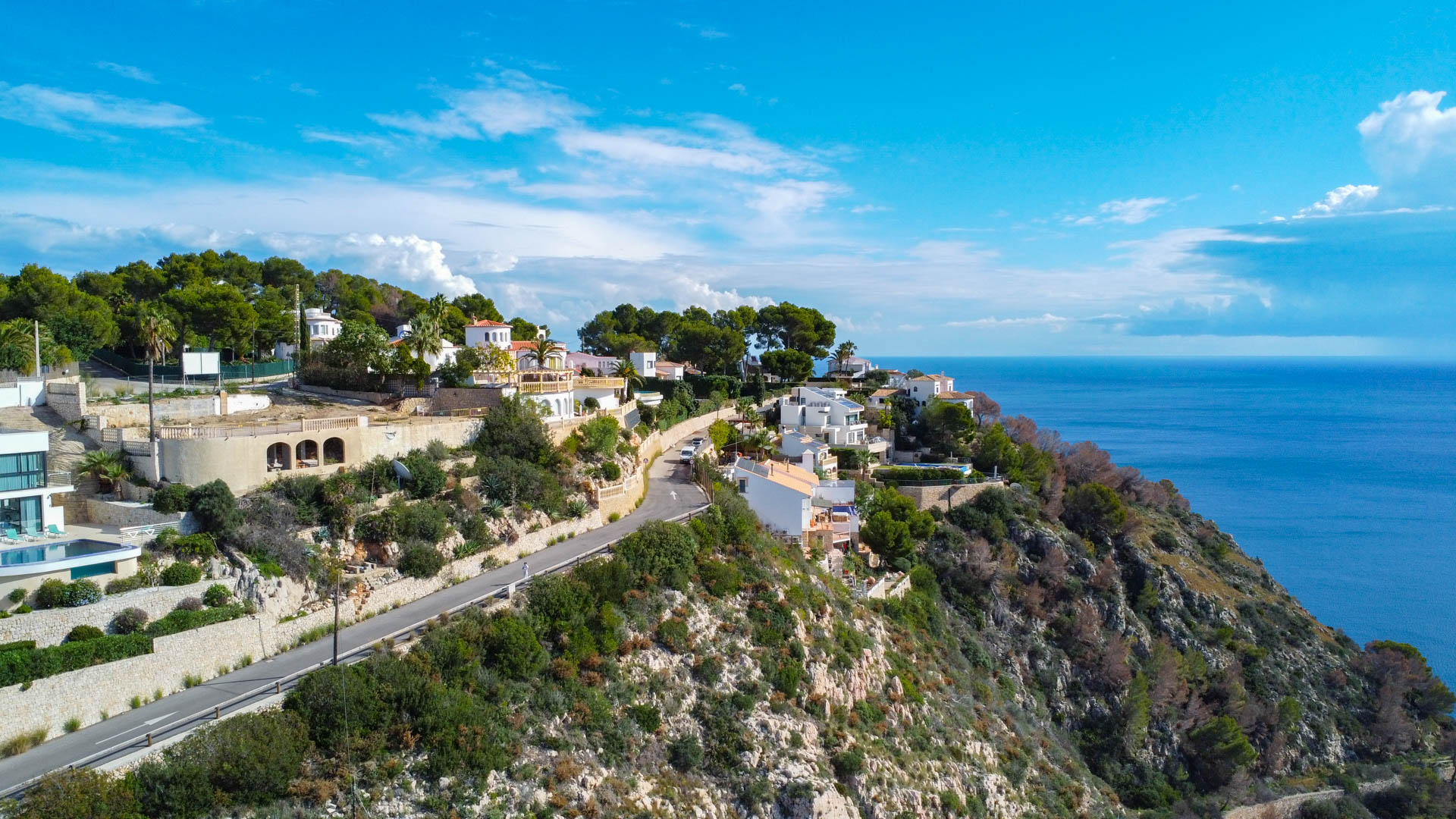 Luftaufnahme des Hanggrundstücks in Granadella mit asphaltierter Zufahrt und weitem Blick auf das Mittelmeer und die zerklüftete Küstenlandschaft.