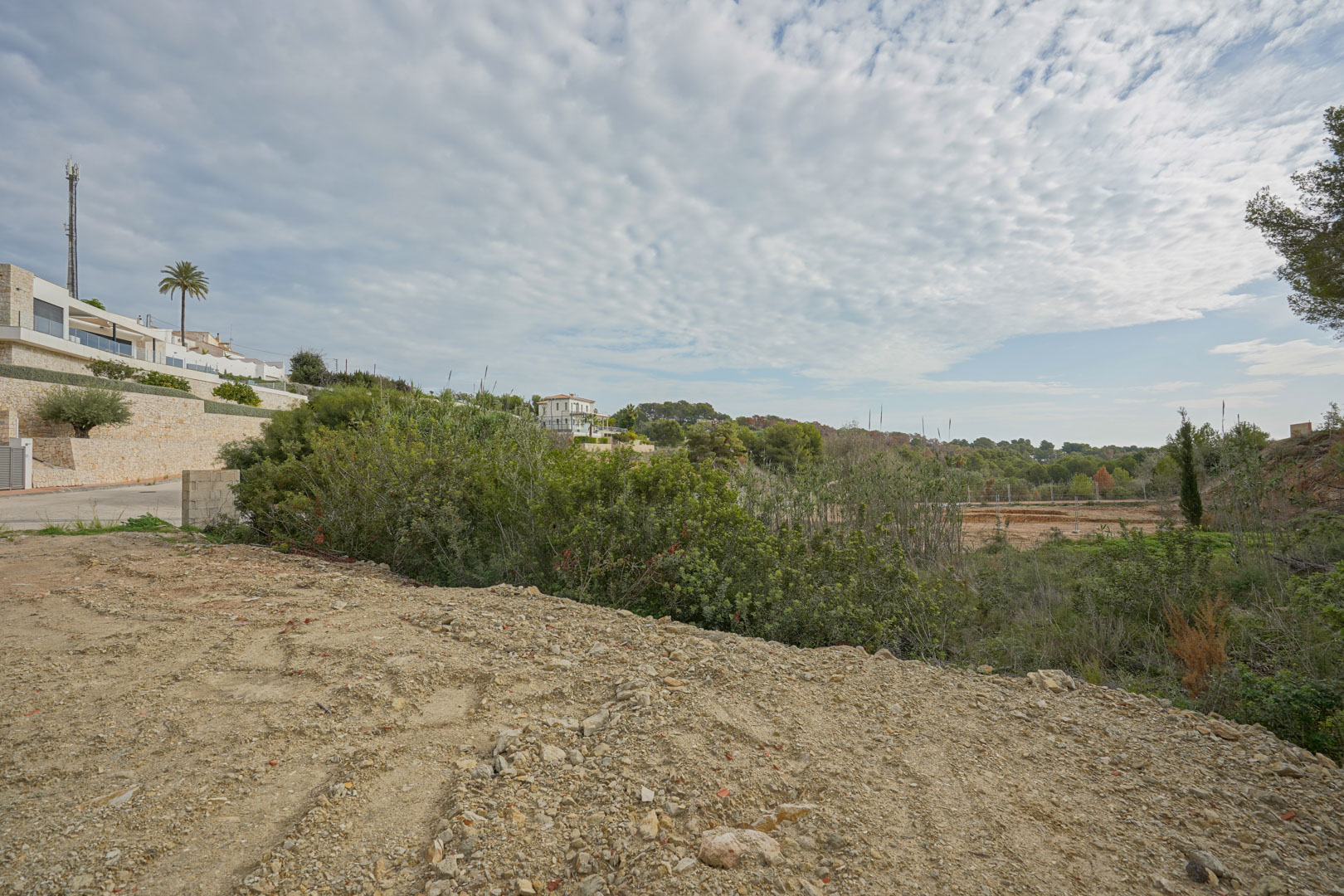 Baureifes Grundstück in einer etablierten Wohngegend, umgeben von mediterraner Vegetation und modernen Villen mit Natursteinmauern.