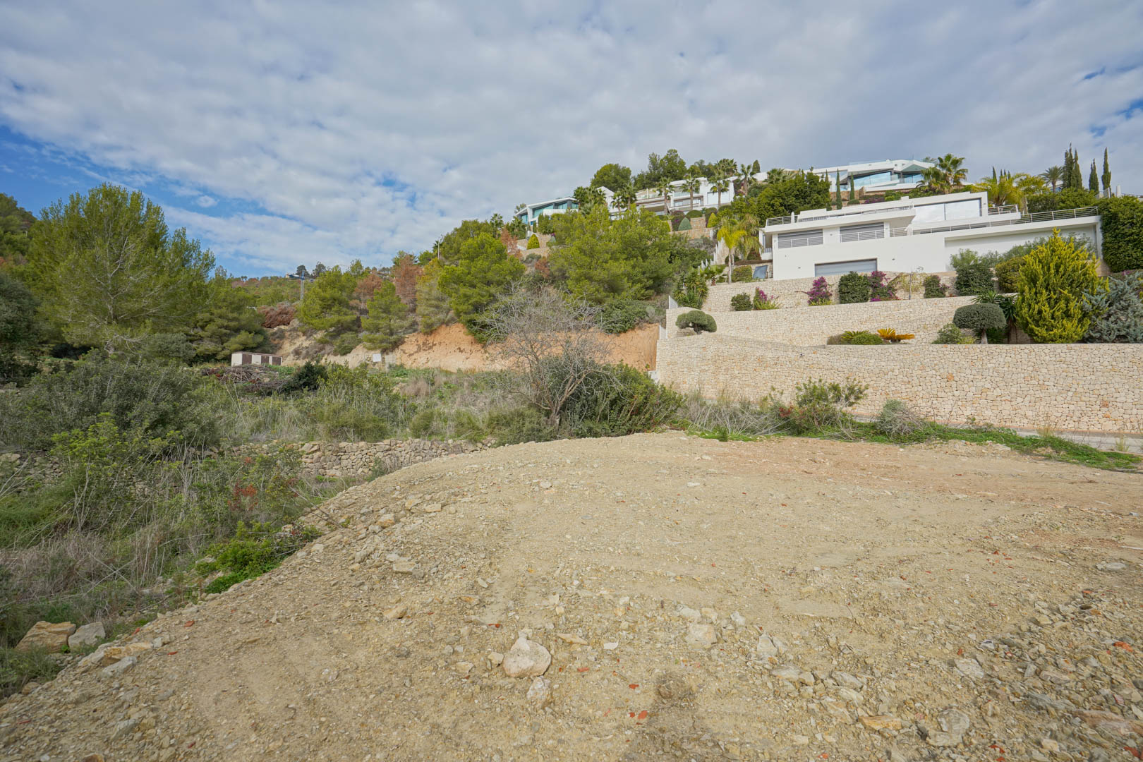 Erschlossenes Baugrundstück in Hanglage mit Blick auf moderne weiße Villen und terrassierte Natursteinmauern inmitten mediterraner Pinienwälder.