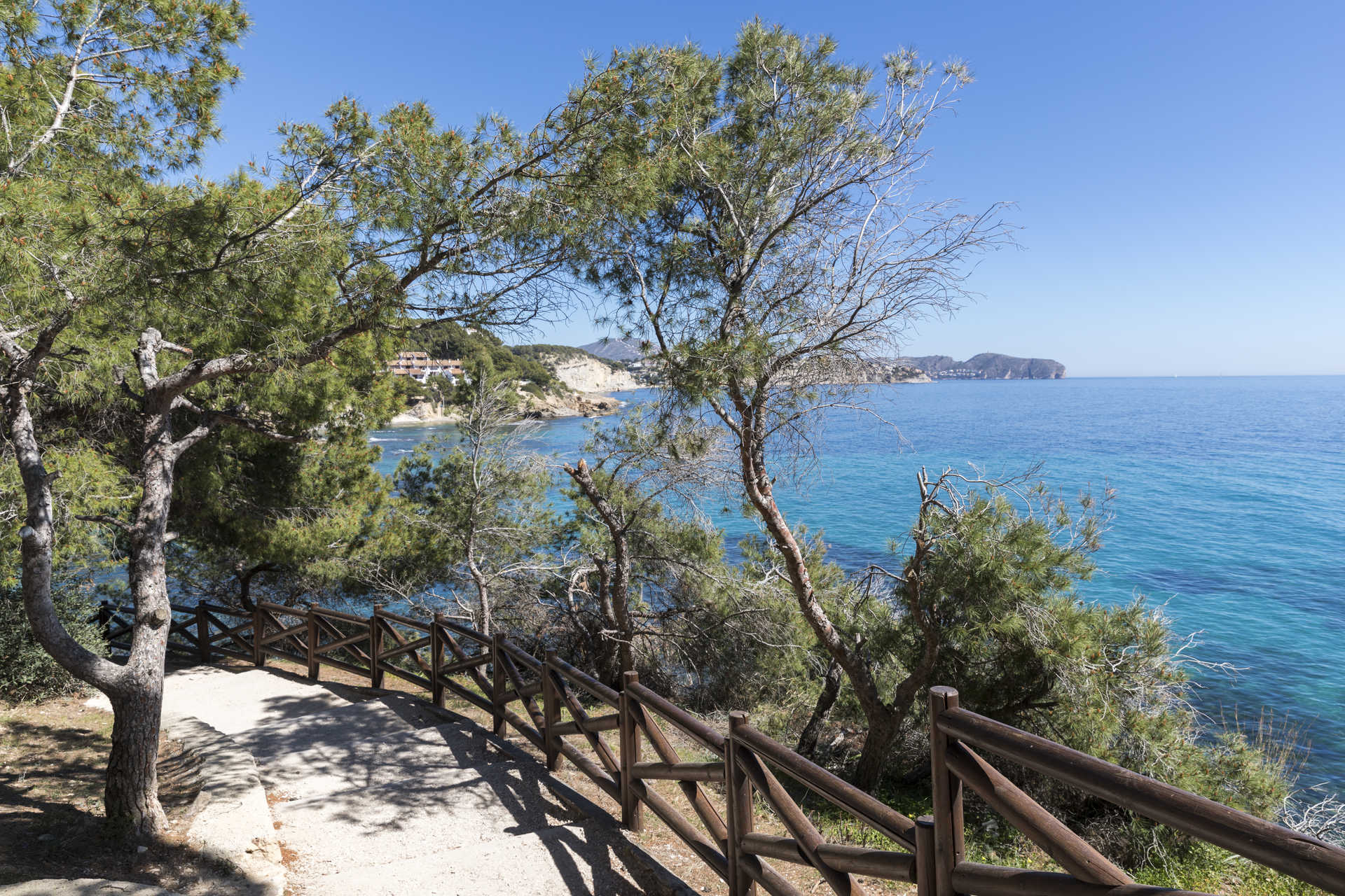 Küstenwanderweg mit Holzgeländer und Pinien, der einen weiten Blick auf das türkisfarbene Mittelmeer in der Nähe des Strandes La Fustera bietet.
