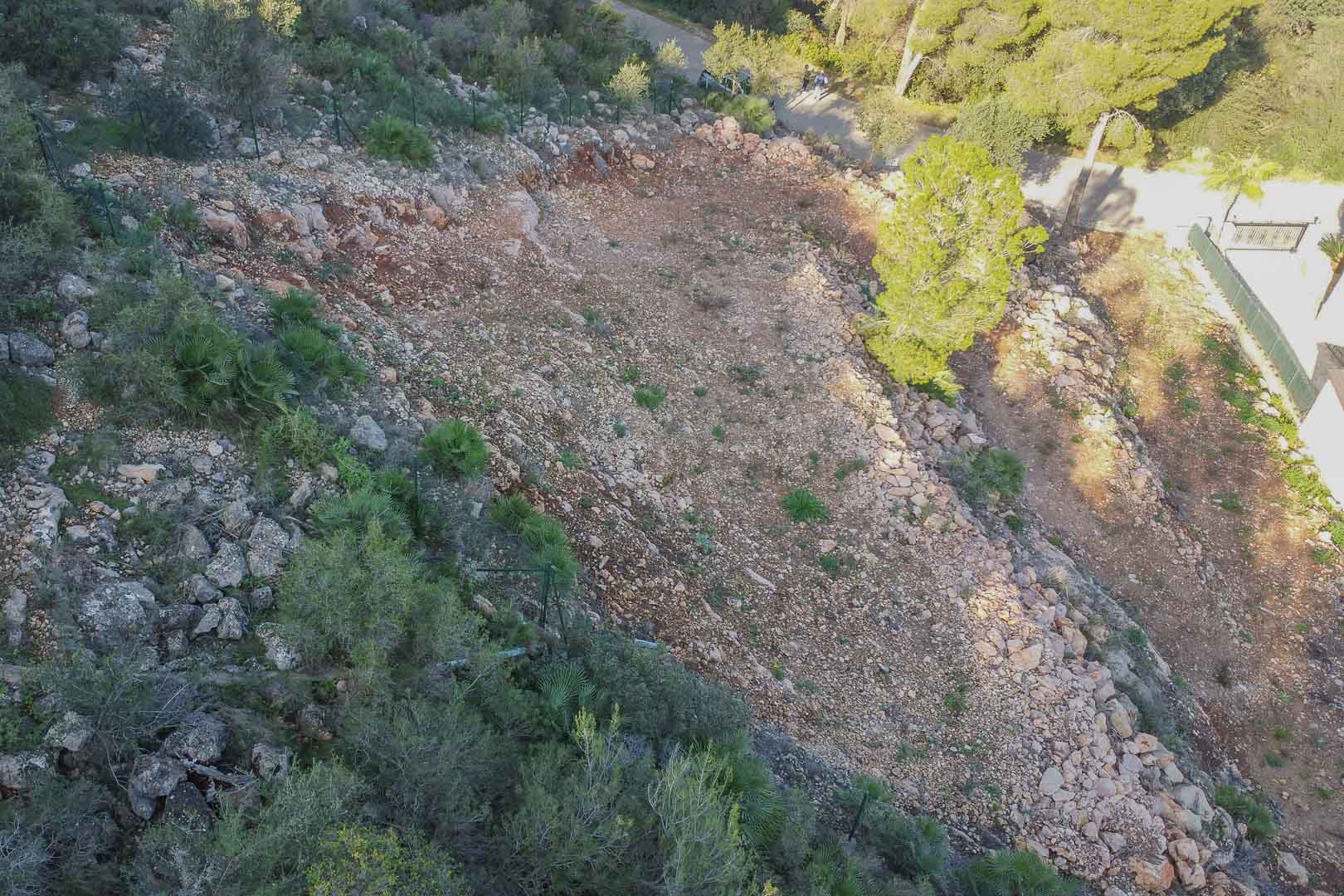 Terrassiertes Baugrundstück in Montepego mit freigelegtem felsigem Boden, Umzäunung und mediterraner Vegetation in unmittelbarer Nähe zur Zufahrtsstraße.