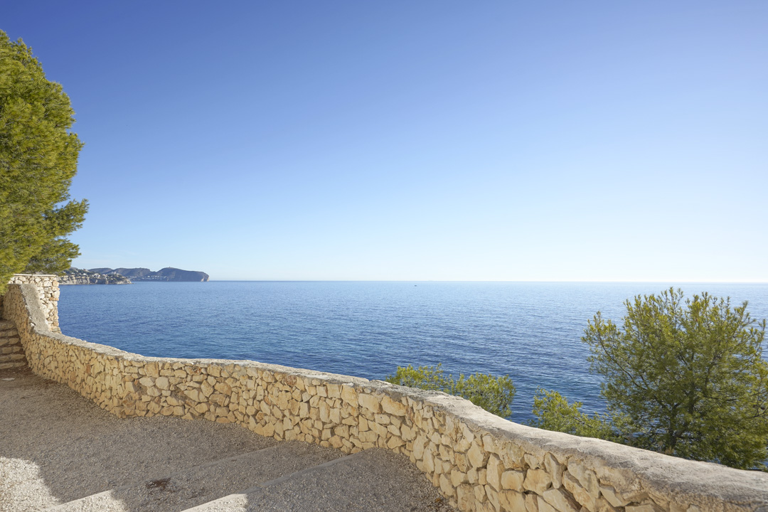 Panoramablick auf das Mittelmeer von einer Terrasse mit Natursteinmauer. Die Szene zeigt einheimische Pinien und einen weiten Blick über die Küste von Calpe.