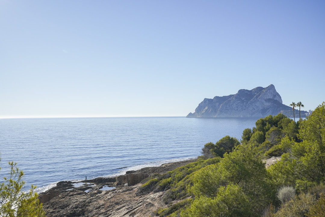 Panoramablick auf den Peñón de Ifach und die Mittelmeerküste mit felsigem Ufer und typisch mediterraner Pinienvegetation unter klarem Himmel.