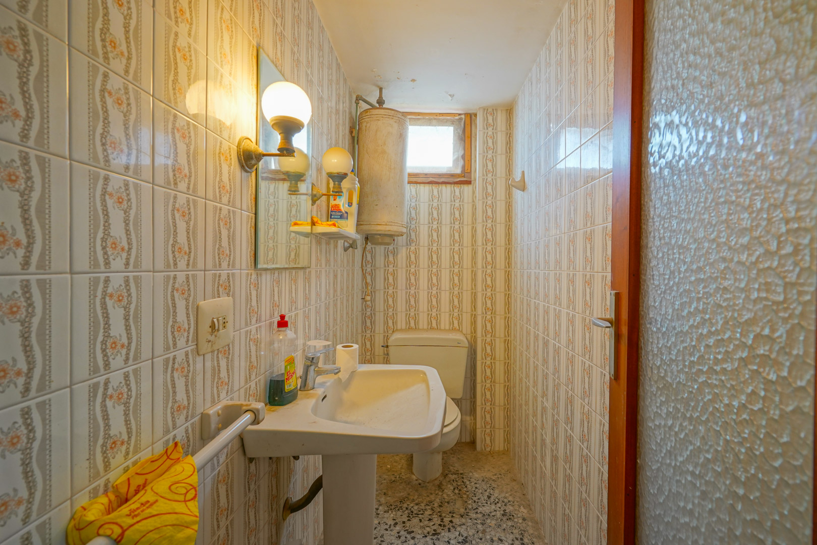 Funktionales Badezimmer mit gemusterten Vintage-Wandfliesen, Standwaschbecken, Elektro-Boiler und originalem Terrazzoboden.