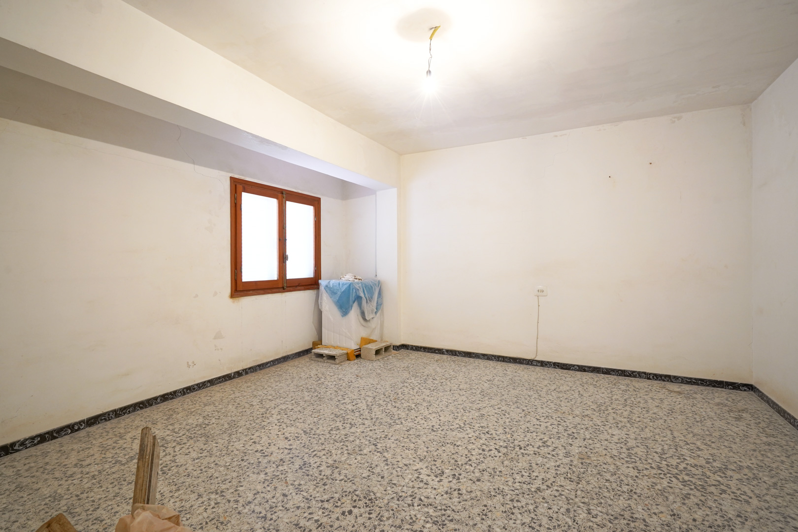 Geräumiges Zimmer mit originalem Terrazzoboden, renovierungsbedürftigen weißen Wänden und einem Holzfenster mit natürlichem Lichteinfall.