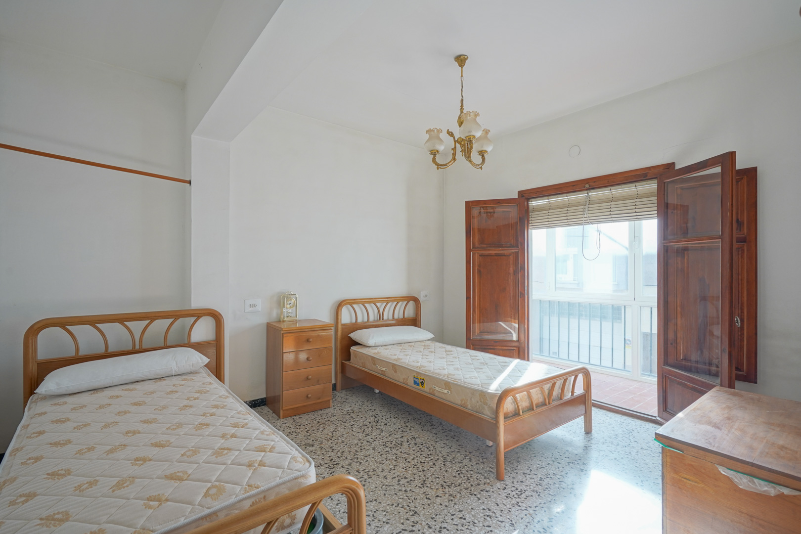 Schlafzimmer mit zwei Einzelbetten, Terrazzoboden und Zugang zum Balkon durch klassische Holzflügeltüren.