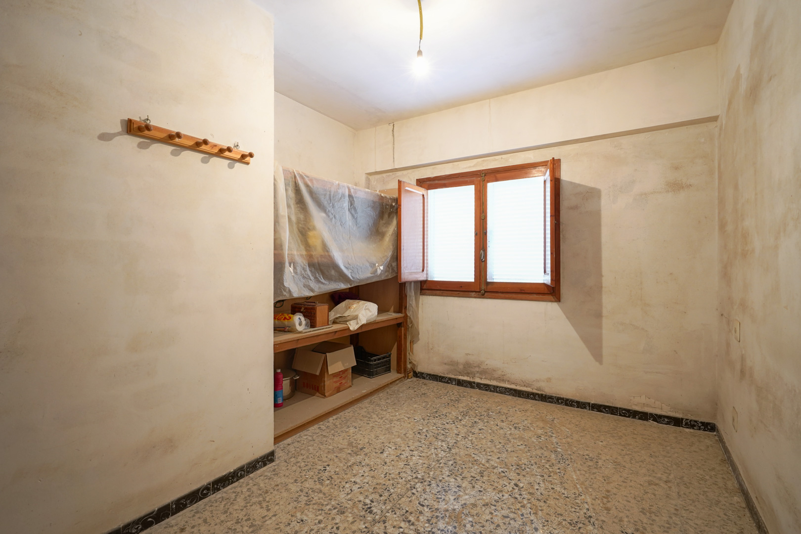 Zimmer mit Terrazzoboden, zweiflügeligem Holzfenster und Einbauregalen, das eine umfassende Renovierung benötigt.