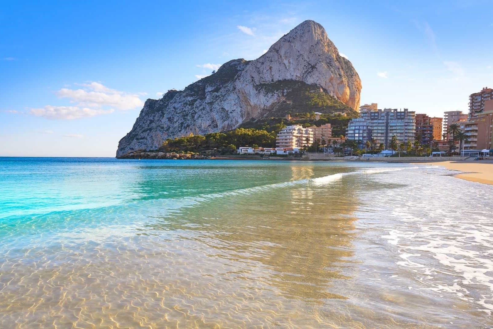 Panoramablick auf den Peñón de Ifach vom Ufer in Calpe, mit kristallklarem türkisfarbenem Wasser und der Skyline der Küstengebäude.