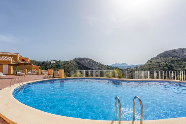 Gemeinschaftlicher Pool in geschwungener Form in Monte Cala Gardens, umgeben von einer Sonnenterrasse mit Liegestühlen und Blick auf die Berge des Naturparks.