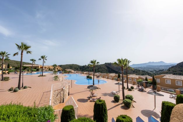 Gemeinschaftsbereich von Monte Cala Gardens mit zwei großen Swimmingpools, weitläufiger Sonnenterrasse mit Palmen und Panoramablick auf die Berge.