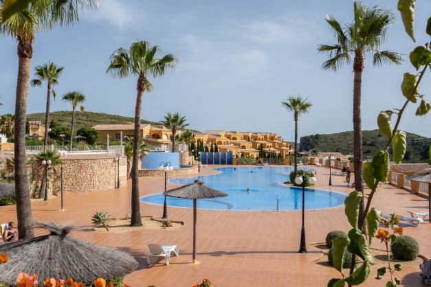 Großer Gemeinschaftspool mit blauen Fliesen, umgeben von einer weitläufigen Terrakotta-Terrasse, Palmen und Natursteinmauern in Monte Cala Gardens.