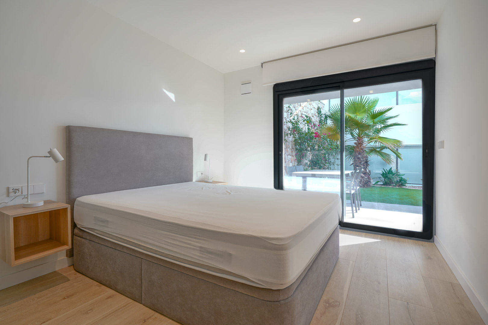 Hauptschlafzimmer mit direktem Zugang zum Garten durch Glasschiebetüren, hellem Holzboden und moderner, minimalistischer Einrichtung mit Einbaustrahlern.