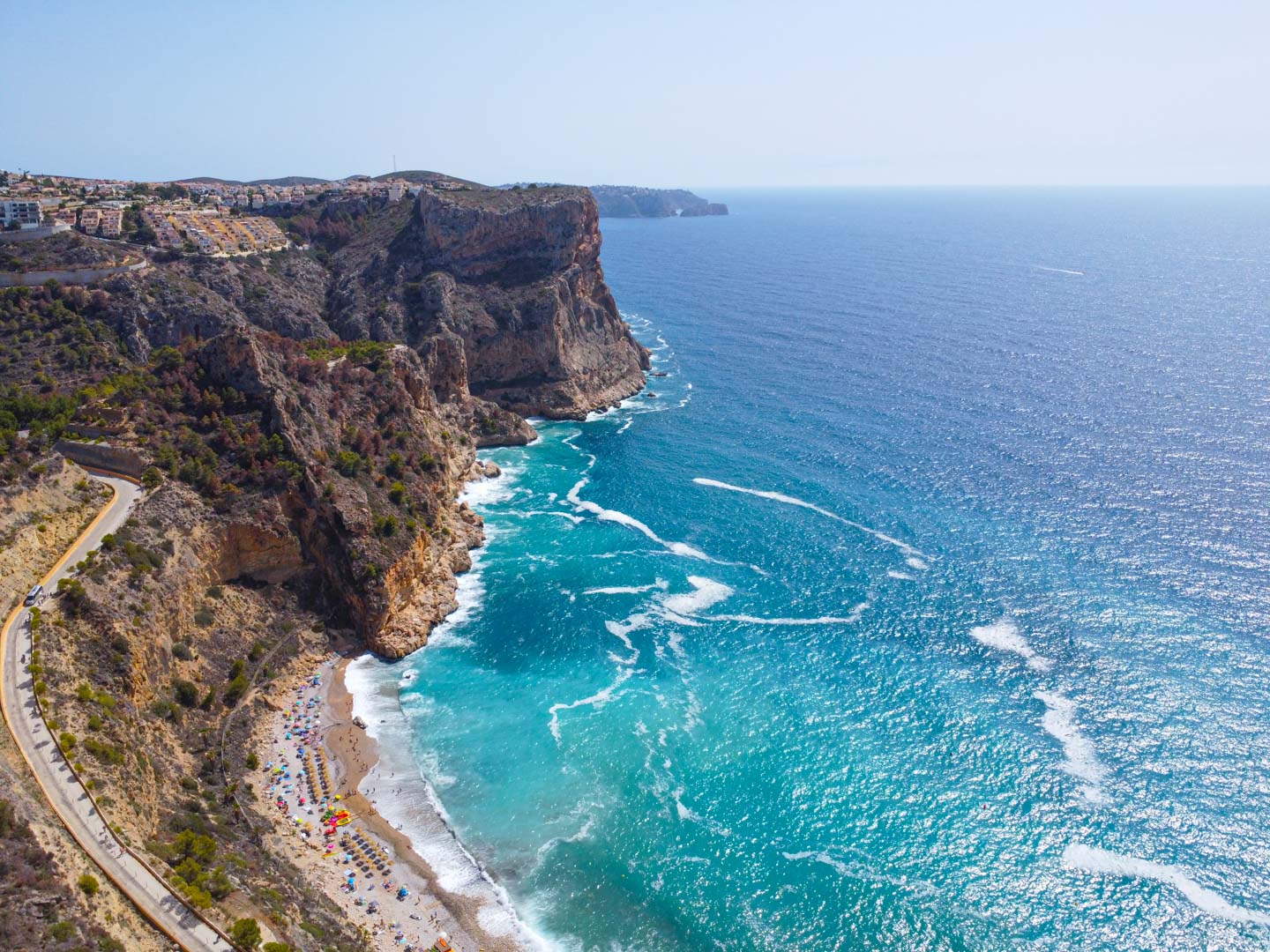 Luftaufnahme der Cala del Moraig in Cumbre del Sol mit markanten Kalksteinfelsen, türkisblauem Wasser und der kurvenreichen Straße zum Strand.