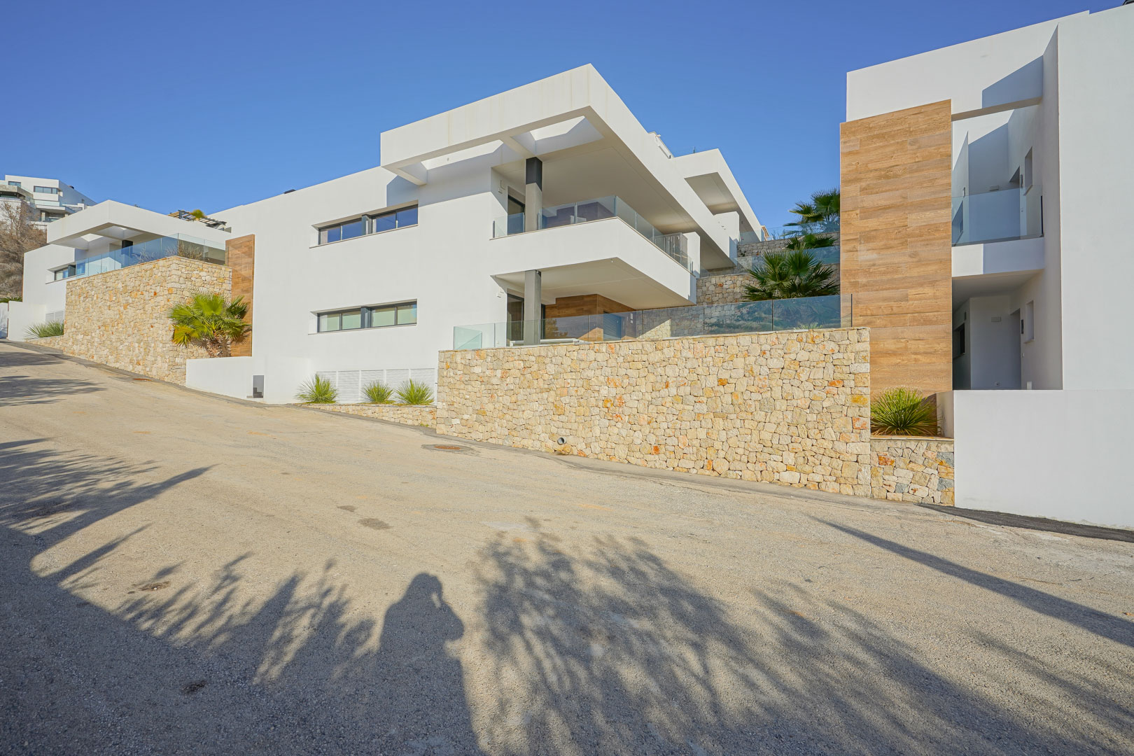 Moderne Fassade in Monte Cala Gardens mit Natursteinmauern, großzügigen Glasterrassen und Akzenten in Holzoptik unter strahlend blauem Himmel.