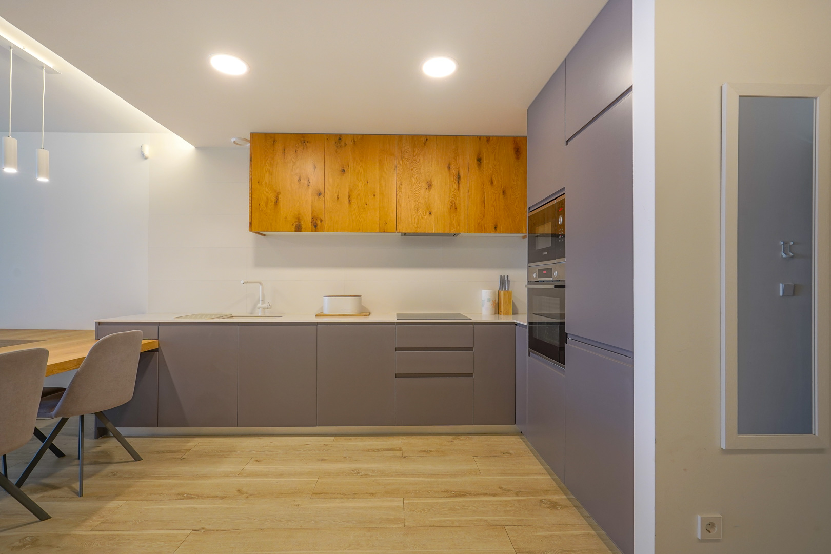 Moderne, minimalistische Küche mit mattgrauen und Echtholz-Schränken, integrierten Einbaugeräten, weißer Arbeitsplatte und hellem Boden in Holzoptik.