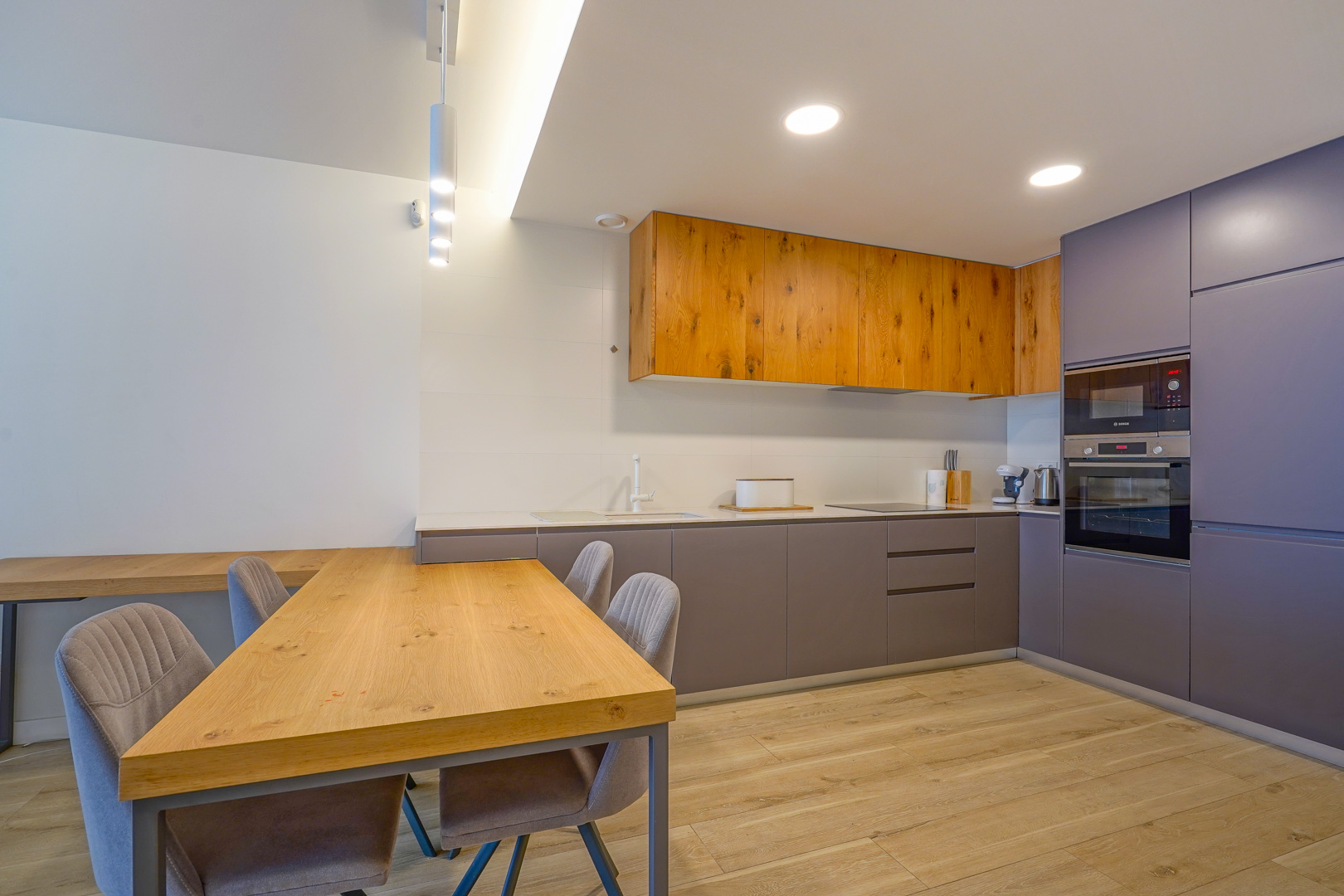 Moderne offene Küche mit Schränken aus Naturholz und Grau, integrierten Geräten und einem passenden Esstisch aus Holz mit Polsterstühlen.