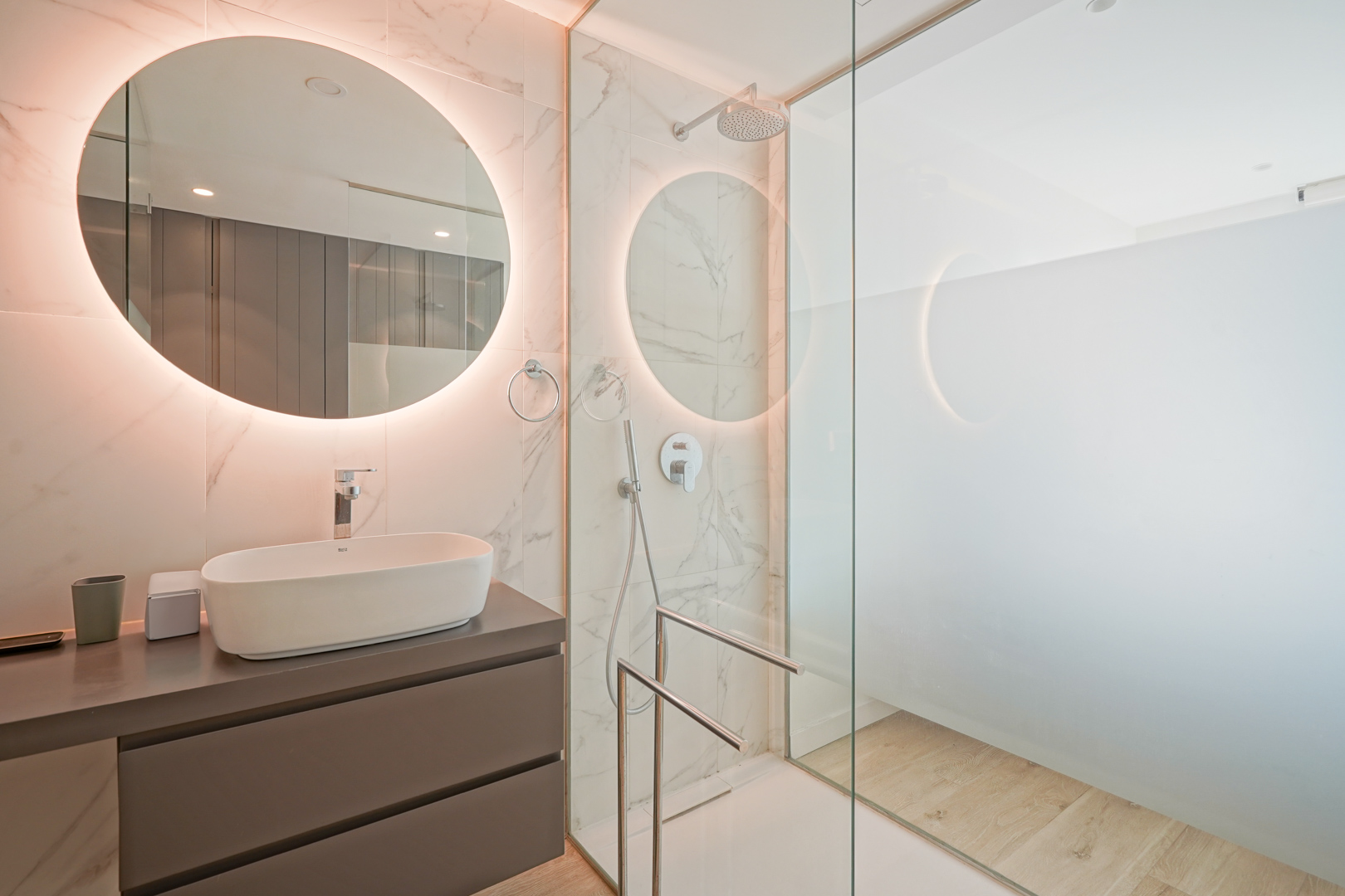Modernes Badezimmer mit Aufsatzwaschbecken, hintergrundbeleuchtetem Rundspiegel und ebenerdiger Dusche mit Glastrennwand und Fliesen in Marmoroptik.