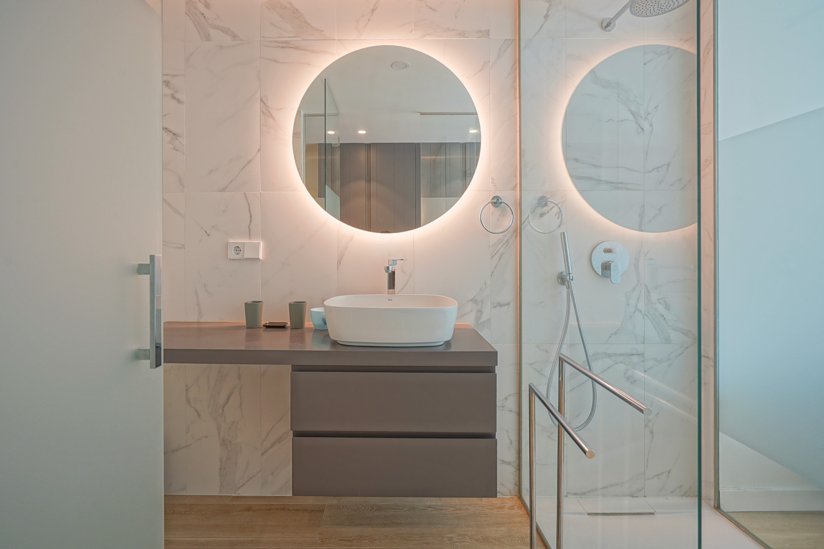 Modernes Badezimmer mit Aufsatzwaschbecken, hinterleuchtetem Rundspiegel und Wandfliesen in Marmoroptik sowie einer Glasdusche.