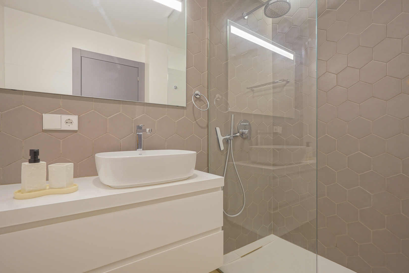 Modernes Badezimmer mit taupefarbenen Sechseckfliesen, Aufsatzwaschbecken auf weißem Unterschrank und ebenerdiger Dusche mit Glaswand.