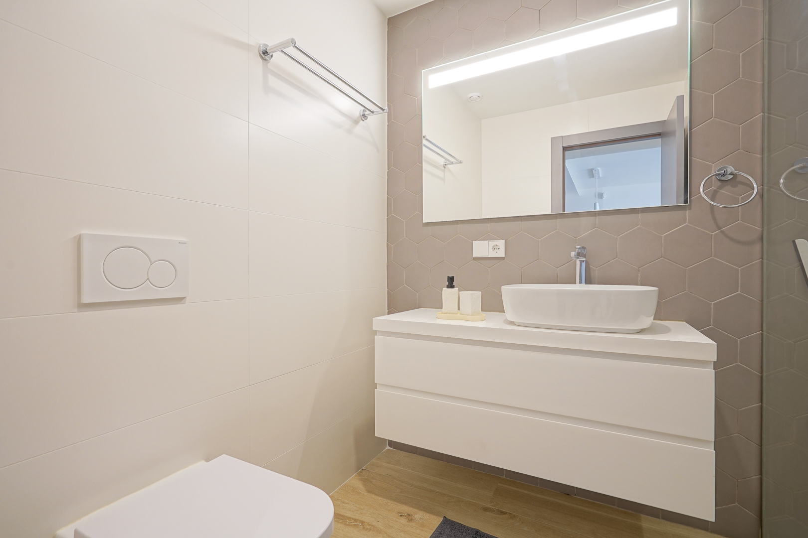 Modernes Badezimmer mit taupefarbenen Sechskantfliesen, weißem Hängeschrank mit Aufsatzwaschbecken und großem Spiegel mit integrierter LED-Beleuchtung.