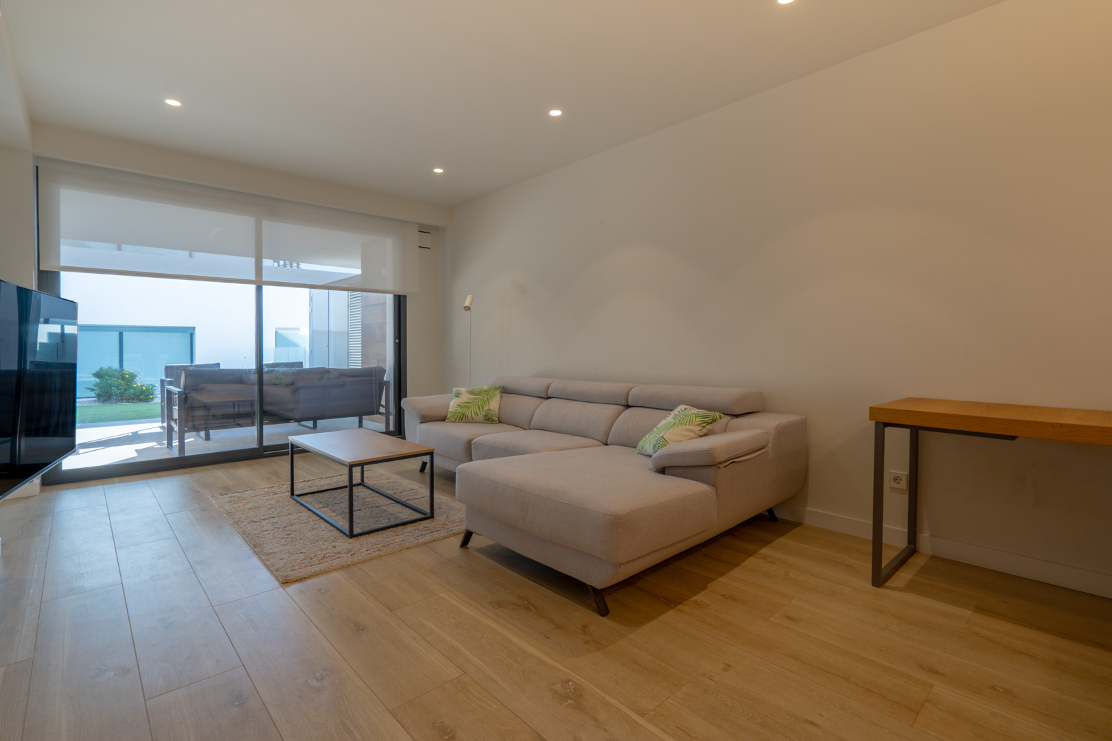 Modernes Wohnzimmer mit grauem Ecksofa, hellem Laminatboden und großen Glasschiebetüren, die direkt auf die überdachte Terrasse und den Garten führen.