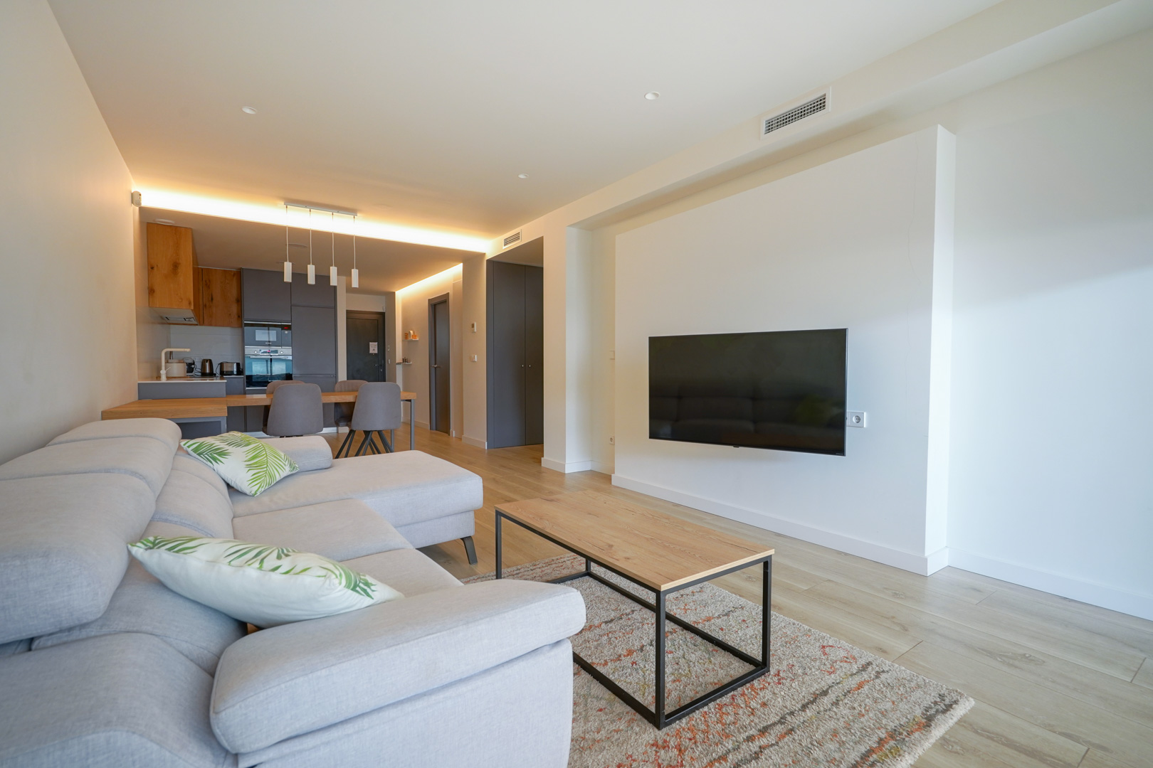 Offener Wohnbereich mit wandmontiertem TV, hellem Holzboden und integrierter moderner Küche mit indirekter Beleuchtung und Holzdetails.