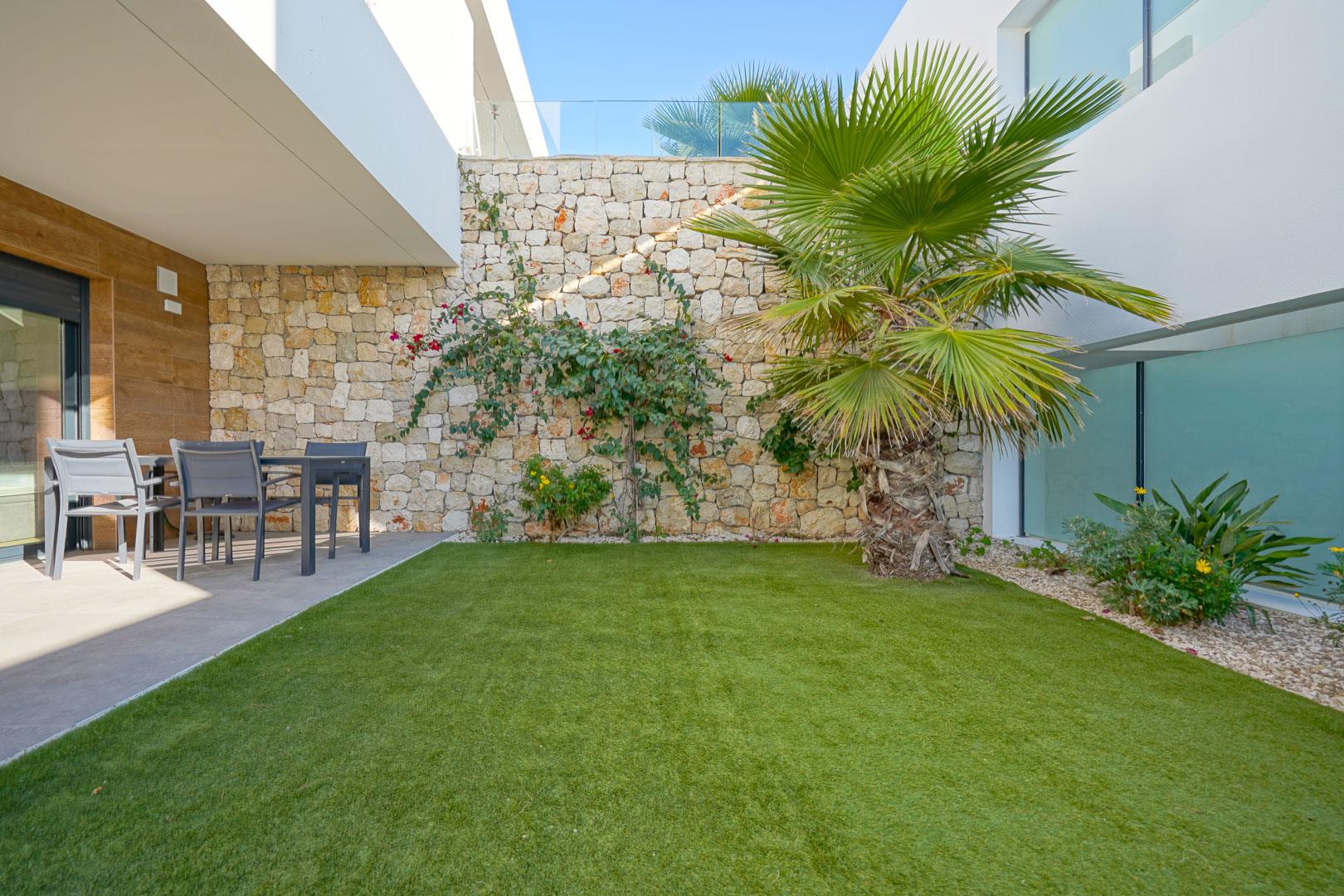 Privater Garten mit Kunstrasen, einer Palme und einer Natursteinmauer. Ein überdachter Terrassenbereich bietet einen schattigen Essplatz im Freien.