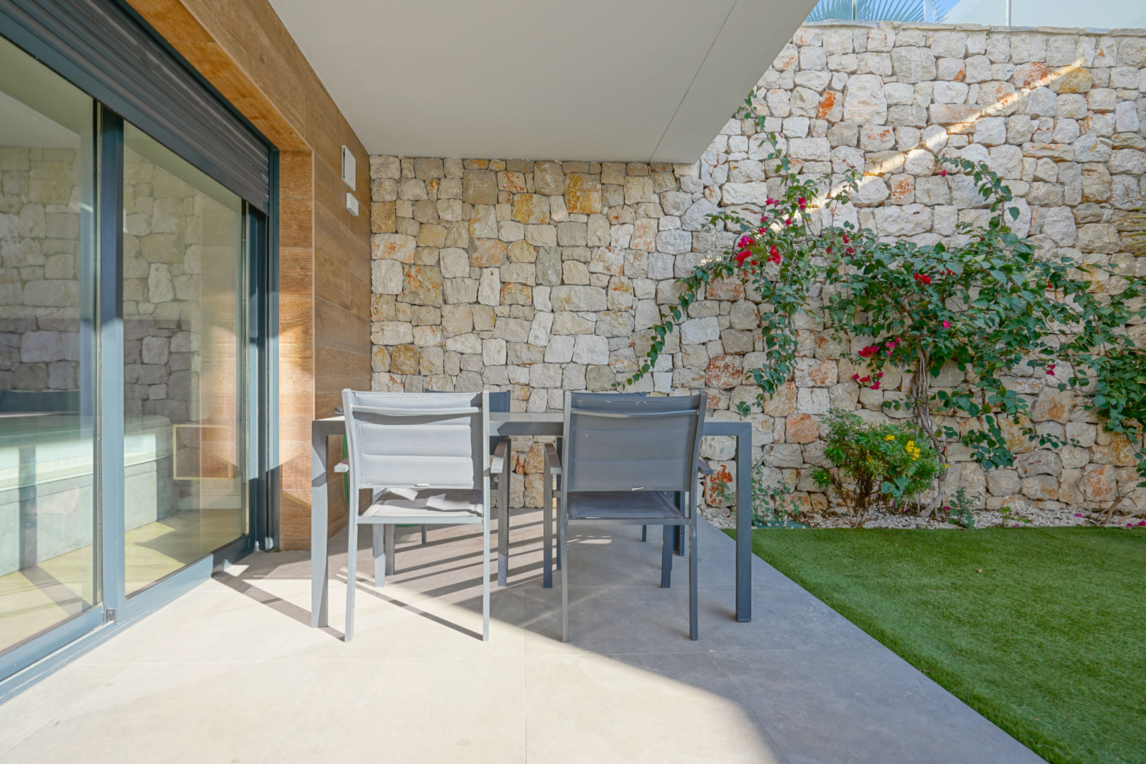 Überdachte Terrasse mit Essplatz im Freien, eingefasst von Natursteinmauern und einem privaten Garten mit Kunstrasen.