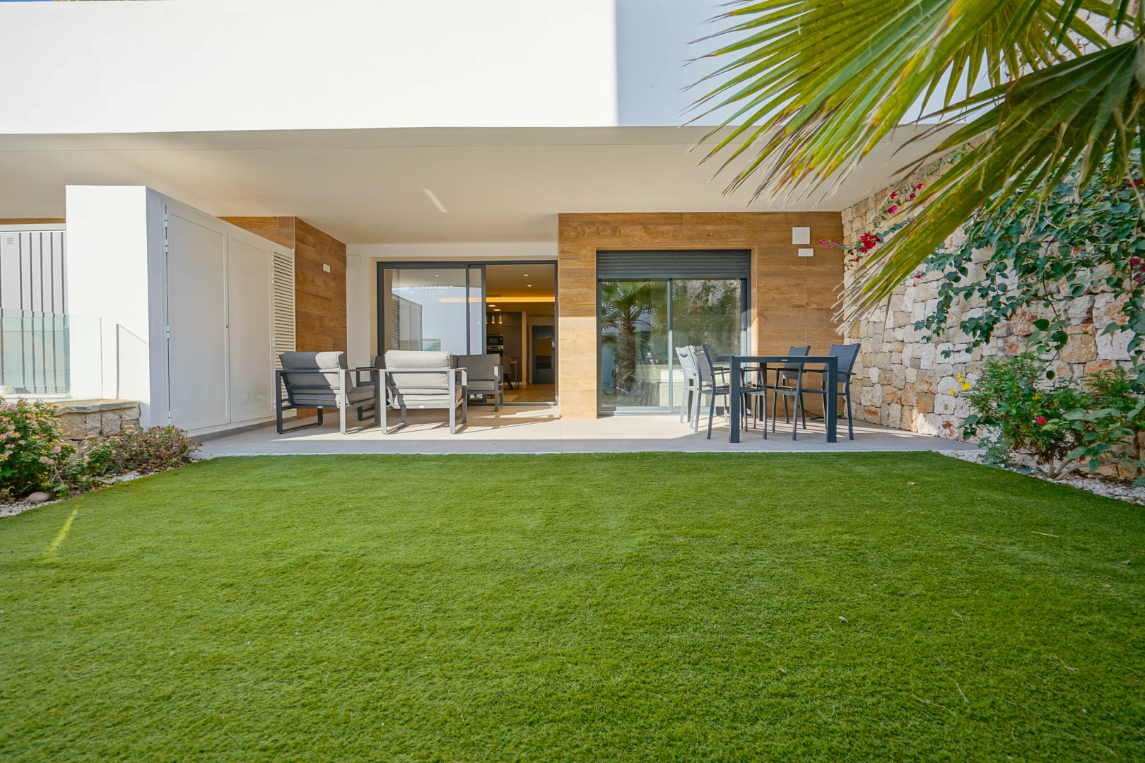 Überdachte Terrasse mit Lounge- und Essbereich, angrenzend an einen privaten Garten mit Rasenfläche und einer Mauer aus Naturstein.