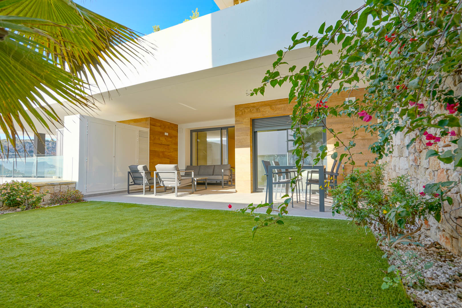 Überdachte Terrasse mit modernen Gartenmöbeln und privatem Garten mit Kunstrasen, begrenzt durch eine Natursteinmauer und mediterrane Pflanzen.