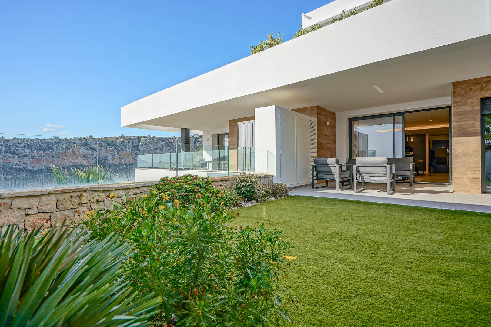 Überdachte Terrasse mit modernen Gartenmöbeln und privatem Rasenanteil, begrenzt durch eine Natursteinmauer mit Blick auf den Naturpark.