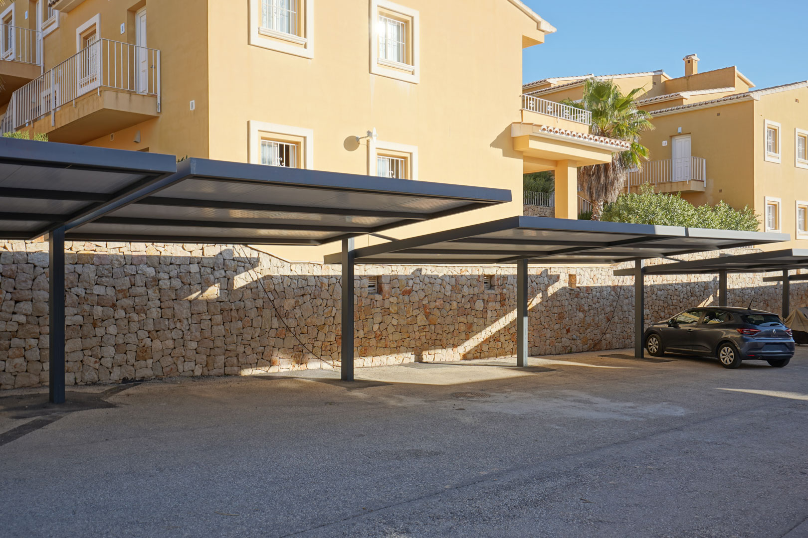 Überdachter Parkplatz mit modernen Metallstrukturen und einer Natursteinmauer, direkt vor dem Wohngebäude gelegen.