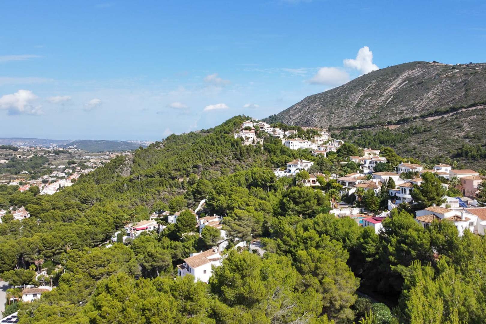 Luftaufnahme des Wohngebiets Paichi in Moraira mit weißen Villen inmitten eines dichten Pinienwaldes und weitem Blick auf die umliegenden Berge.