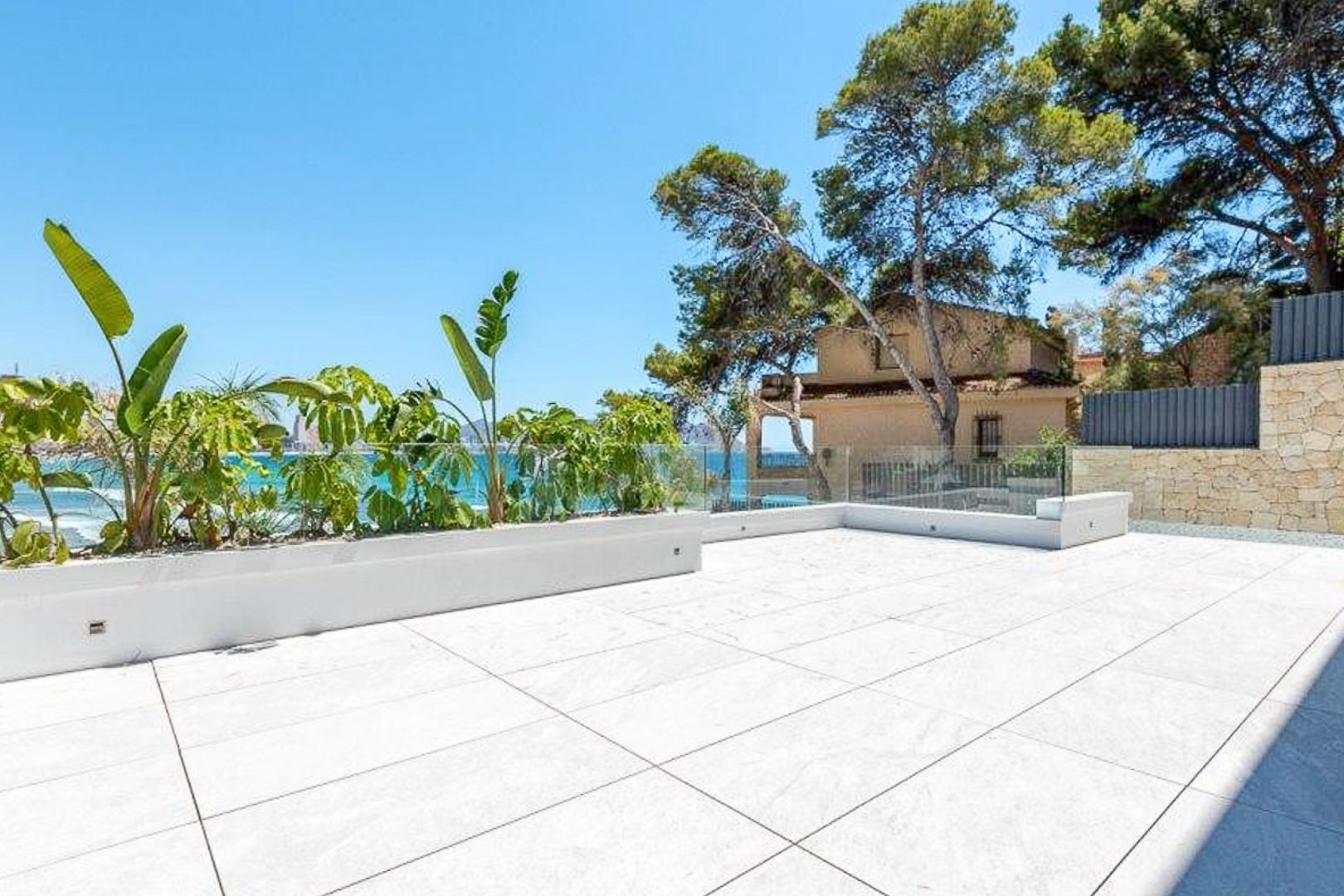 Außen-Terrasse mit großformatigen Feinsteinzeugfliesen, rahmenlosem Glasgeländer und integrierten Pflanzgefäßen mit Blick auf das Mittelmeer.