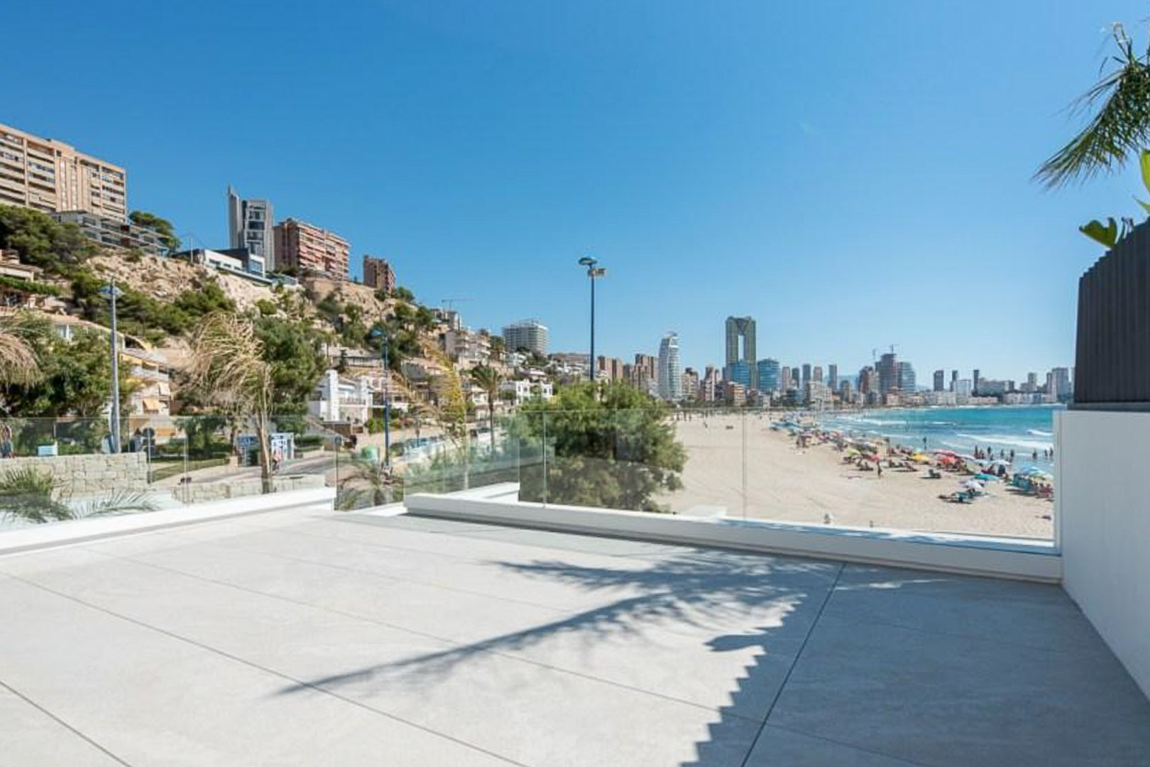 Außen-Terrasse mit großformatigen Steinplatten und Glasgeländer, die einen Panoramablick auf den Sandstrand und die Skyline von Benidorm bietet.