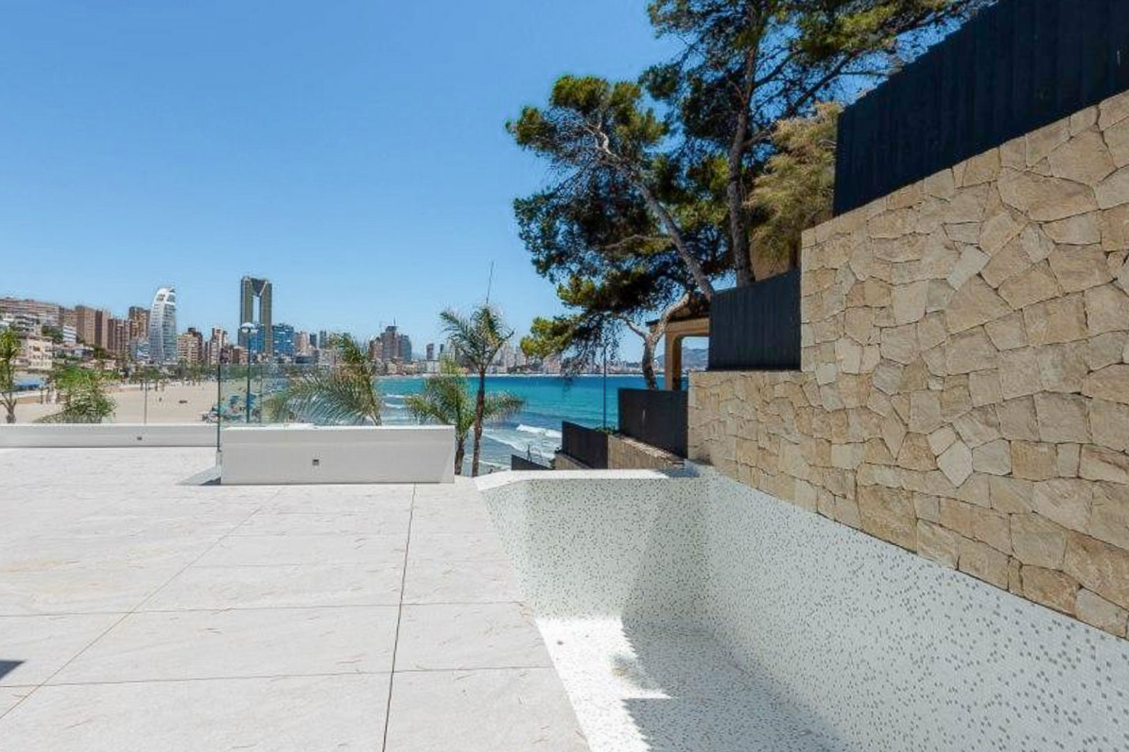 Außen-Terrasse mit hellen Steinplatten und weißem Mosaikpool, die einen Panoramablick auf das Meer und die Skyline von Benidorm neben einer Natursteinmauer bietet.