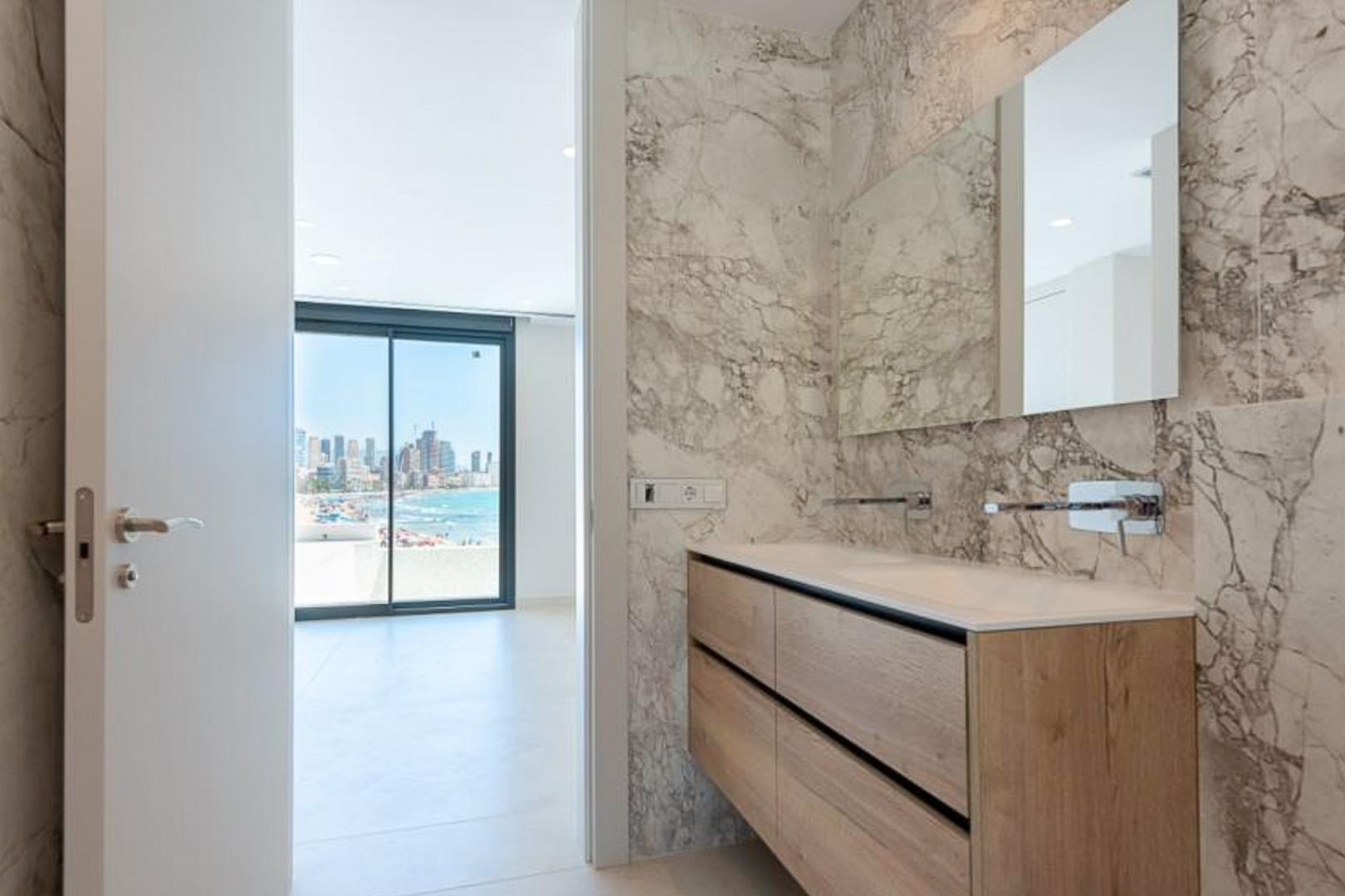 Elegantes Badezimmer mit Marmorwänden, Doppelwaschtisch in Holzoptik und Unterputzarmaturen mit Blick auf die Küste von Benidorm.