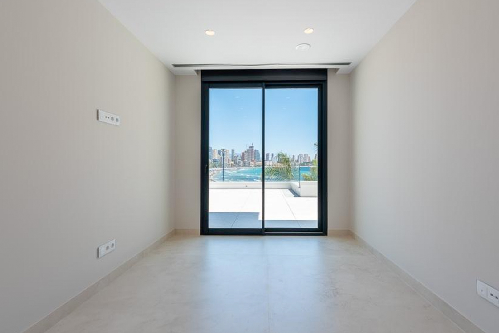 Heller Raum mit Feinsteinzeugboden, Einbaustrahlern und großen Glasschiebetüren, die auf eine Terrasse mit Blick auf das Meer und die Skyline von Benidorm führen.