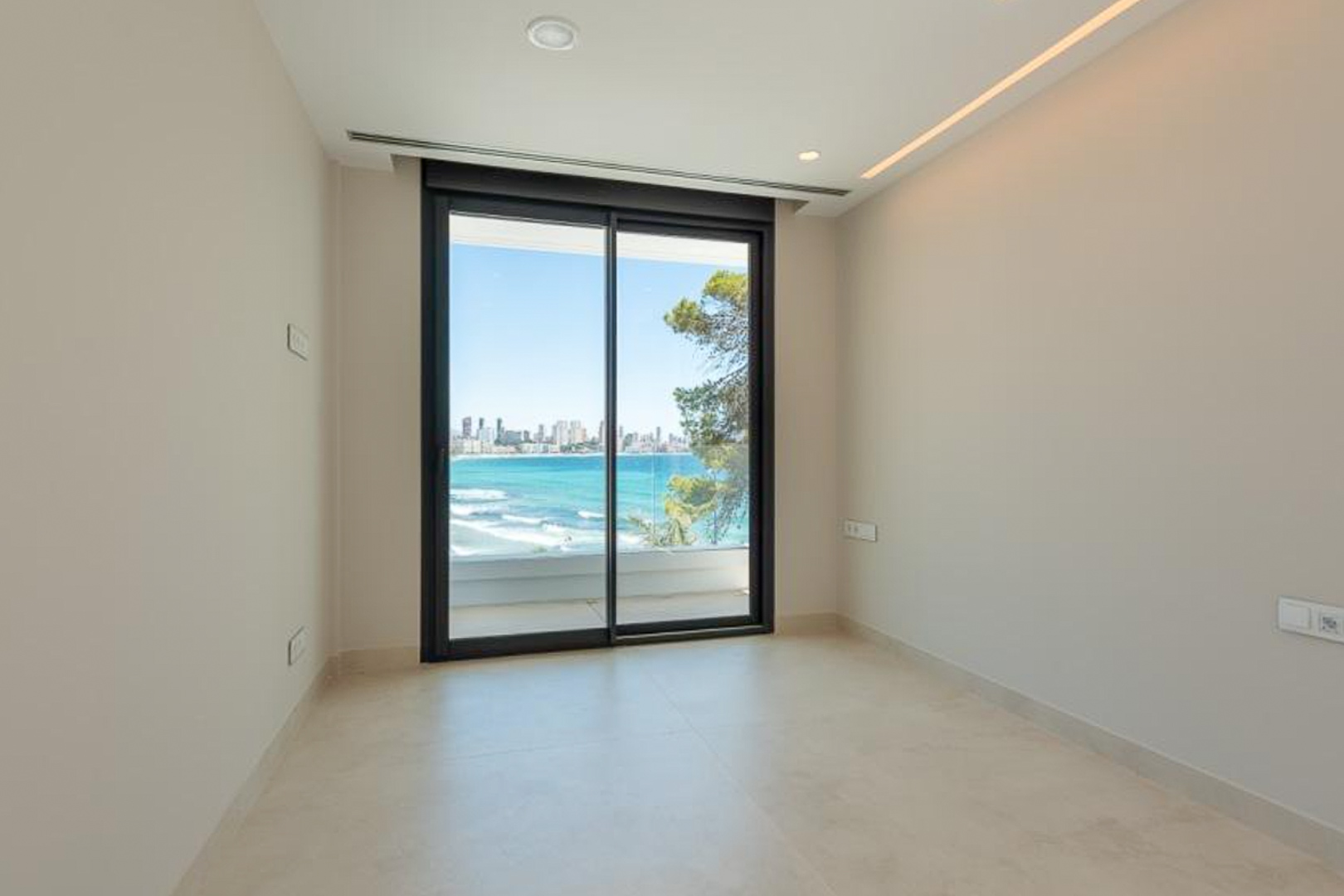 Heller Raum mit Feinsteinzeugboden, integrierter LED-Beleuchtung und Glasschiebetüren zur Terrasse mit Panoramablick auf das Meer und Benidorm.