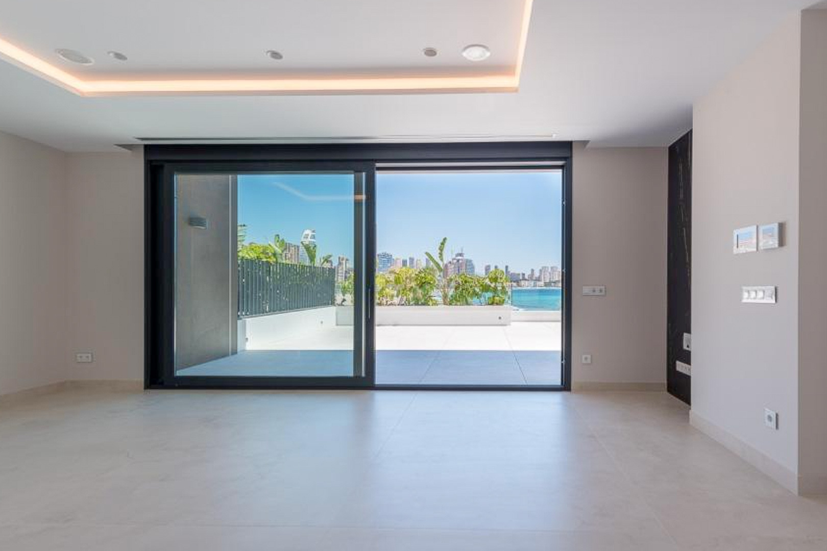 Heller Wohnbereich mit großformatigen Keramikfliesen und Glasschiebetüren, die auf eine private Terrasse mit Panoramablick auf die Skyline von Benidorm führen.