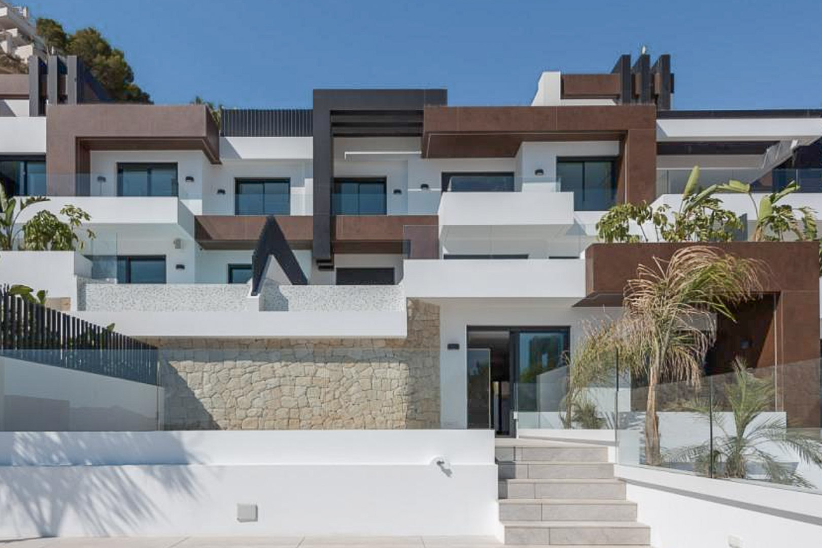 Moderne Architekturfassade mit weißen und braunen geometrischen Formen, Natursteinmauern, Glasgeländern und abgestuften Terrassen mit Palmen.
