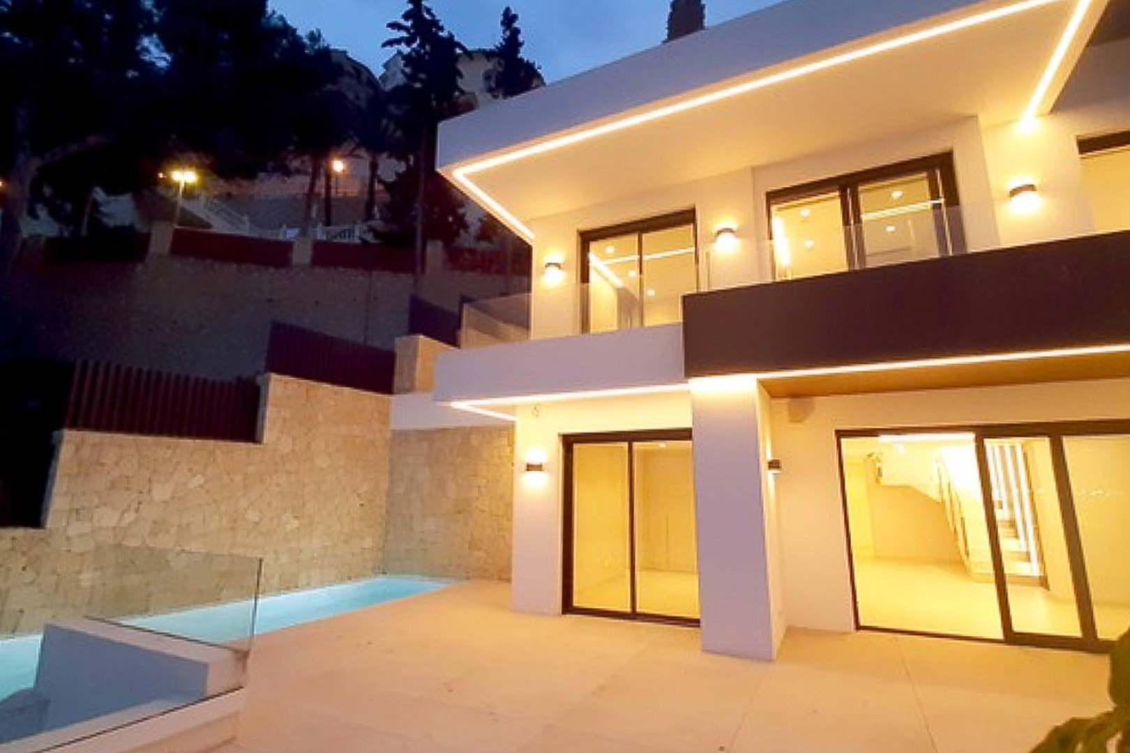 Moderne zweistöckige Fassade mit integrierter LED-Beleuchtung, großen Glasschiebetüren und einer Terrasse mit privatem Pool an einer Natursteinmauer.