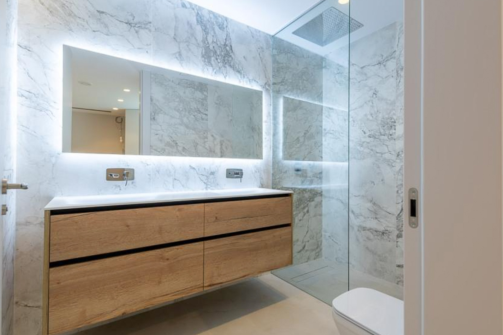 Modernes Badezimmer mit Marmoroptik-Fliesen, schwebendem Doppelwaschtisch aus Holz und hinterleuchtetem Spiegel. Begehbare Dusche mit Glastrennwand.