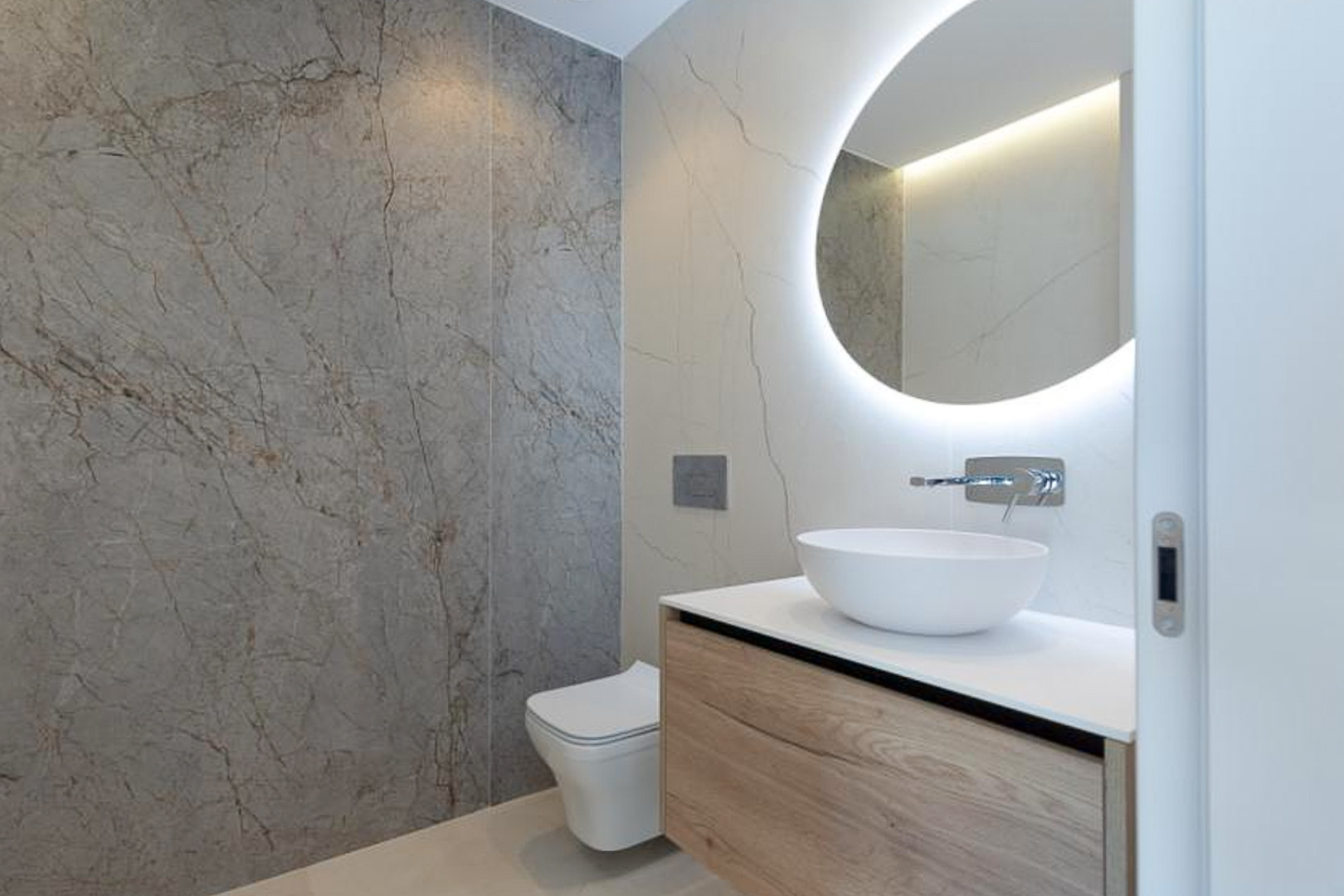 Modernes Badezimmer mit schwebendem Holz-Waschtisch, Aufsatzwaschbecken und hinterleuchtetem Rundspiegel vor großformatigen Fliesen in Marmoroptik.