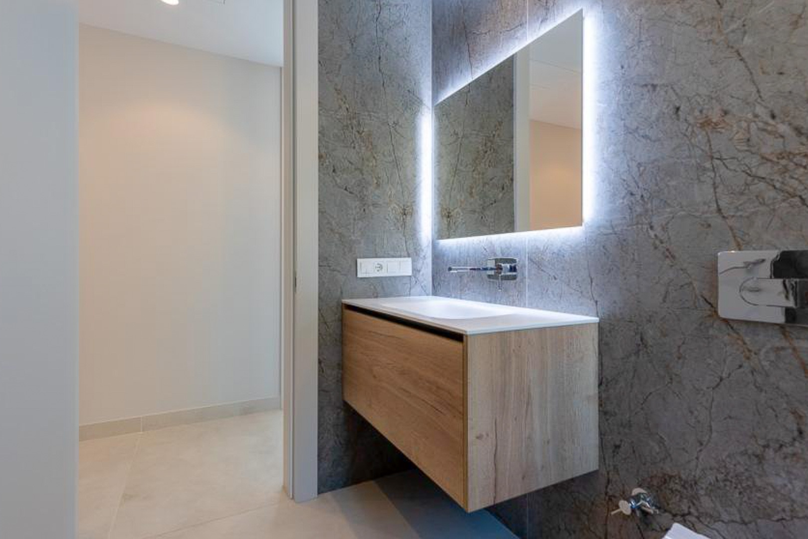 Modernes Gäste-WC mit schwebendem Holzwaschtisch, integriertem Becken und hinterleuchtetem Spiegel vor einer Wand in grauer Marmoroptik.