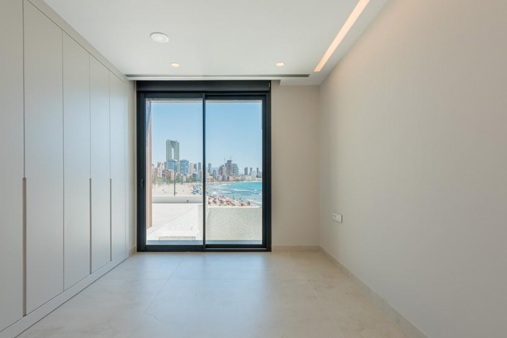 Schlafzimmer mit deckenhohen Einbauschränken, LED-Lichtleiste und Schiebetür mit Blick auf den Poniente-Strand und die Skyline von Benidorm.
