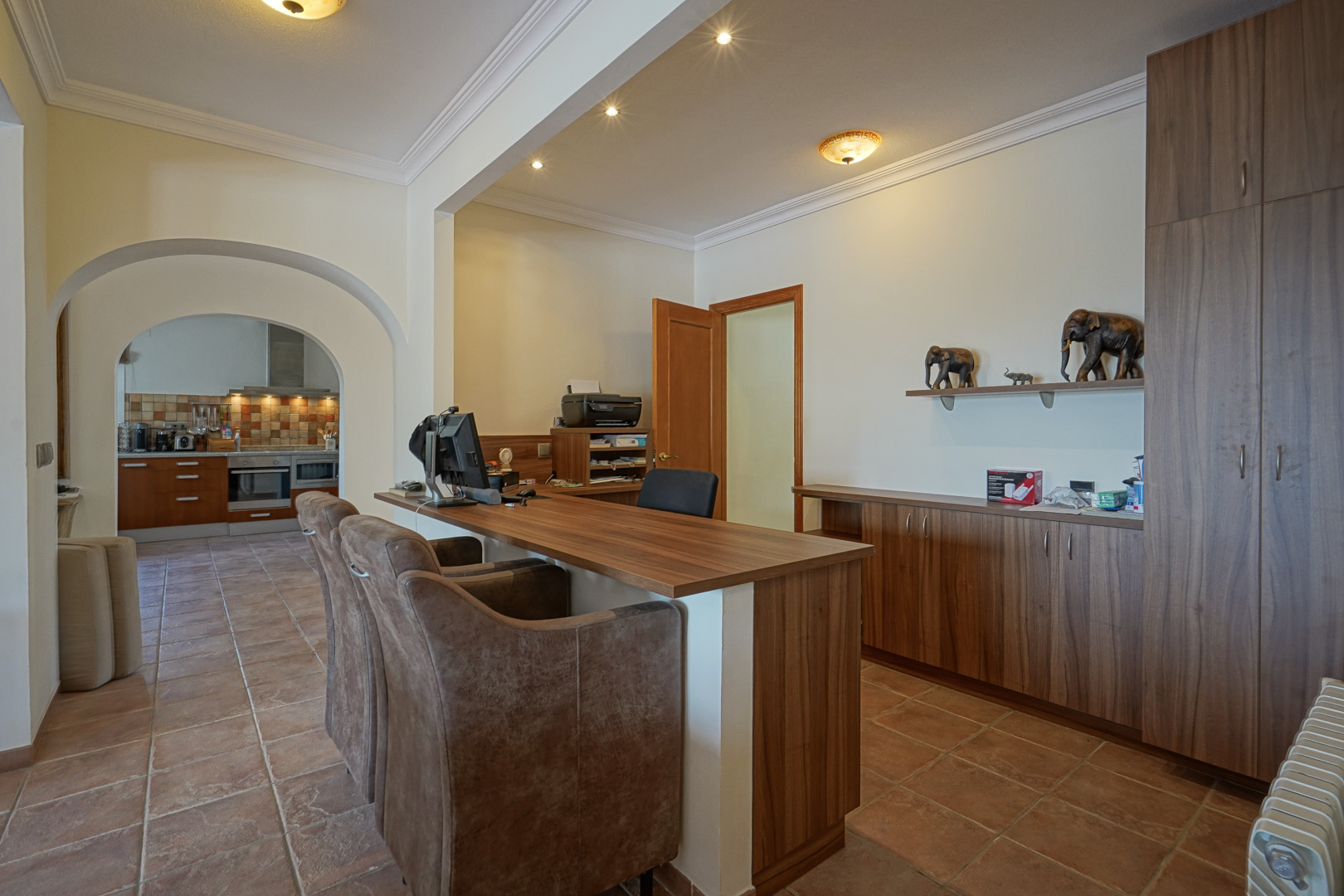 Arbeitsbereich mit maßgefertigten Holzschränken, Terrakottaboden und Rundbögen, die den Blick in die angrenzende Küche freigeben.