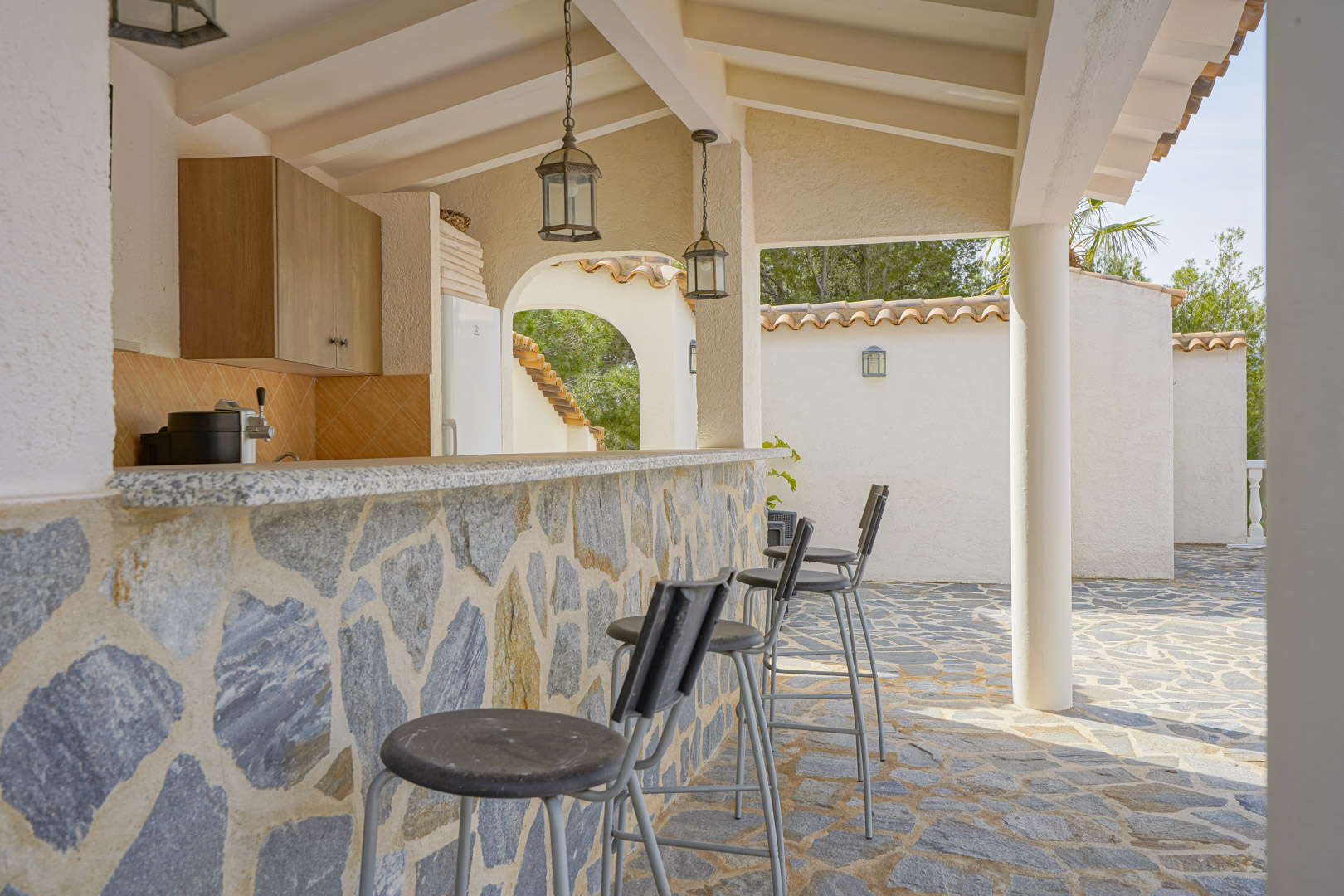 Außenküche mit Natursteinbar, Granitarbeitsplatte und Barhockern unter einer überdachten Terrasse mit Holzbalkendecke und rustikalen Pendelleuchten.